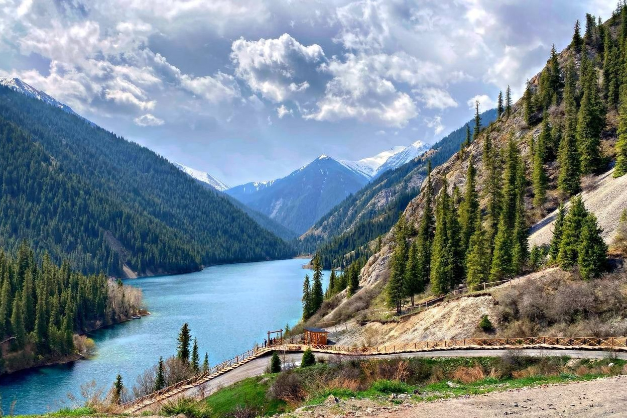 Женщина идет по тропе среди красных скал в пустынной местности. Небо пасмурное, создавая атмосферу спокойствия и уединенности. Вокруг величественные скальные образования Чарынского каньона с характерными слоями песчаника.