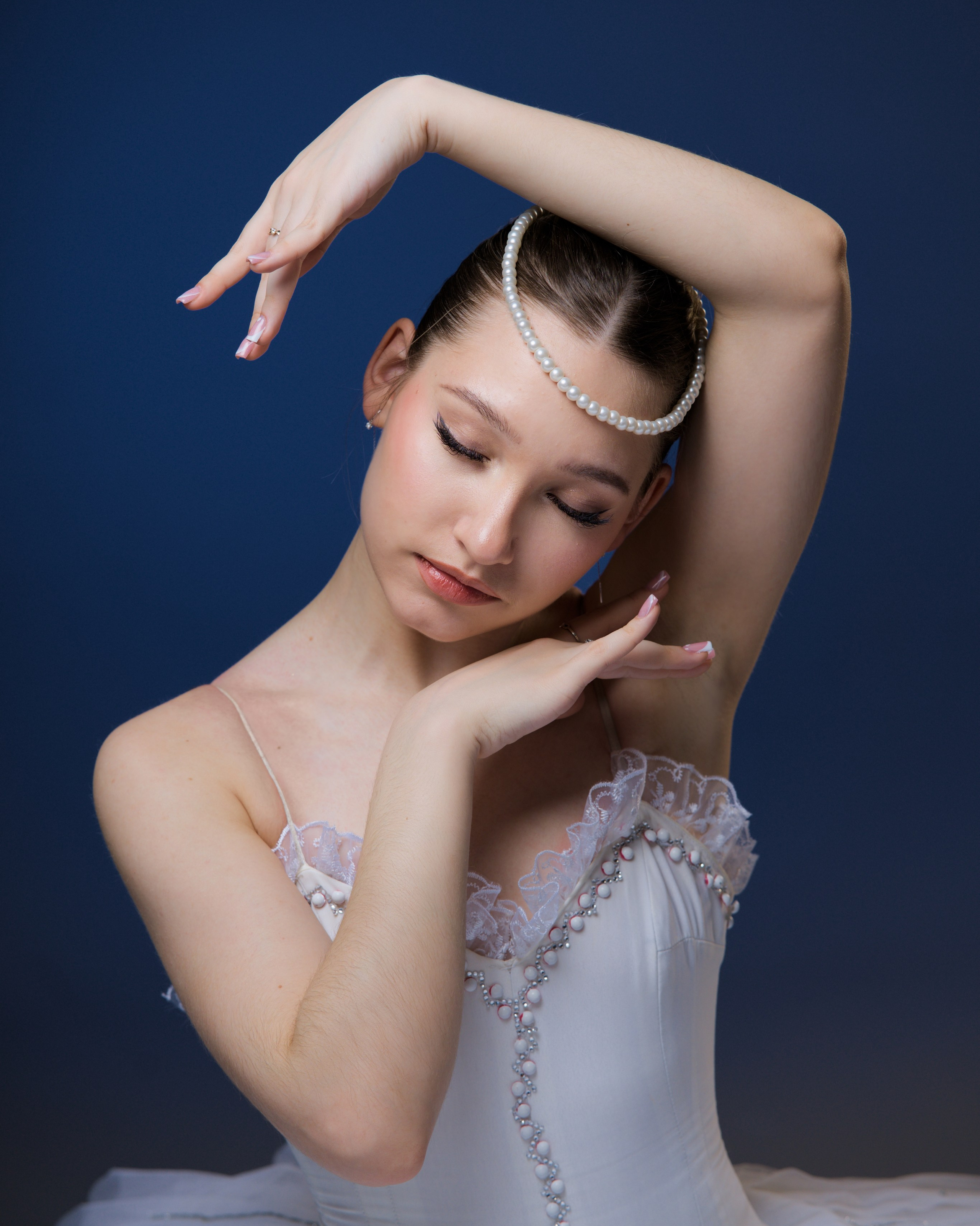Фотосессия для балерины в белой пачке с жемчугом, классическая балетная поза рук, студийный портрет танцовщицы на синем фоне.