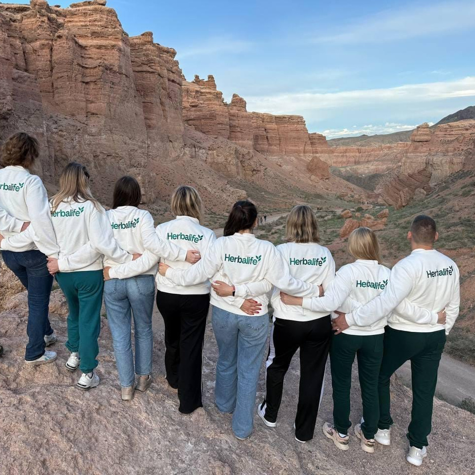 Группа сотрудников, стоящих на улице возле черного автомобиля на фоне ярко окрашенных осенних деревьев. Участники улыбаются и машут руками, создавая дружелюбную и позитивную атмосферу в корпоративном мероприятии. На заднем плане видны здания и зелёные деревья. Яркие цвета осенних листьев подчёркивают радостное настроение сцены.