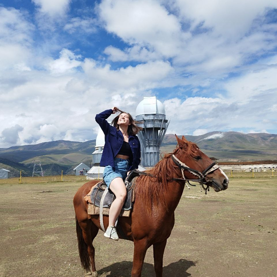 Живописное бирюзовое озеро Иссык рядом с Алматы, окружённое скалами и хвойными деревьями. На переднем плане женщина в бежевом пальто делает фотографию, а другая девушка сидит на скале, любуясь водоёмом. Вдалеке, на левом берегу, ещё один человек отдыхает на камнях. Атмосфера спокойствия и умиротворения, подчёркнутая мягким освещением и отражением воды.