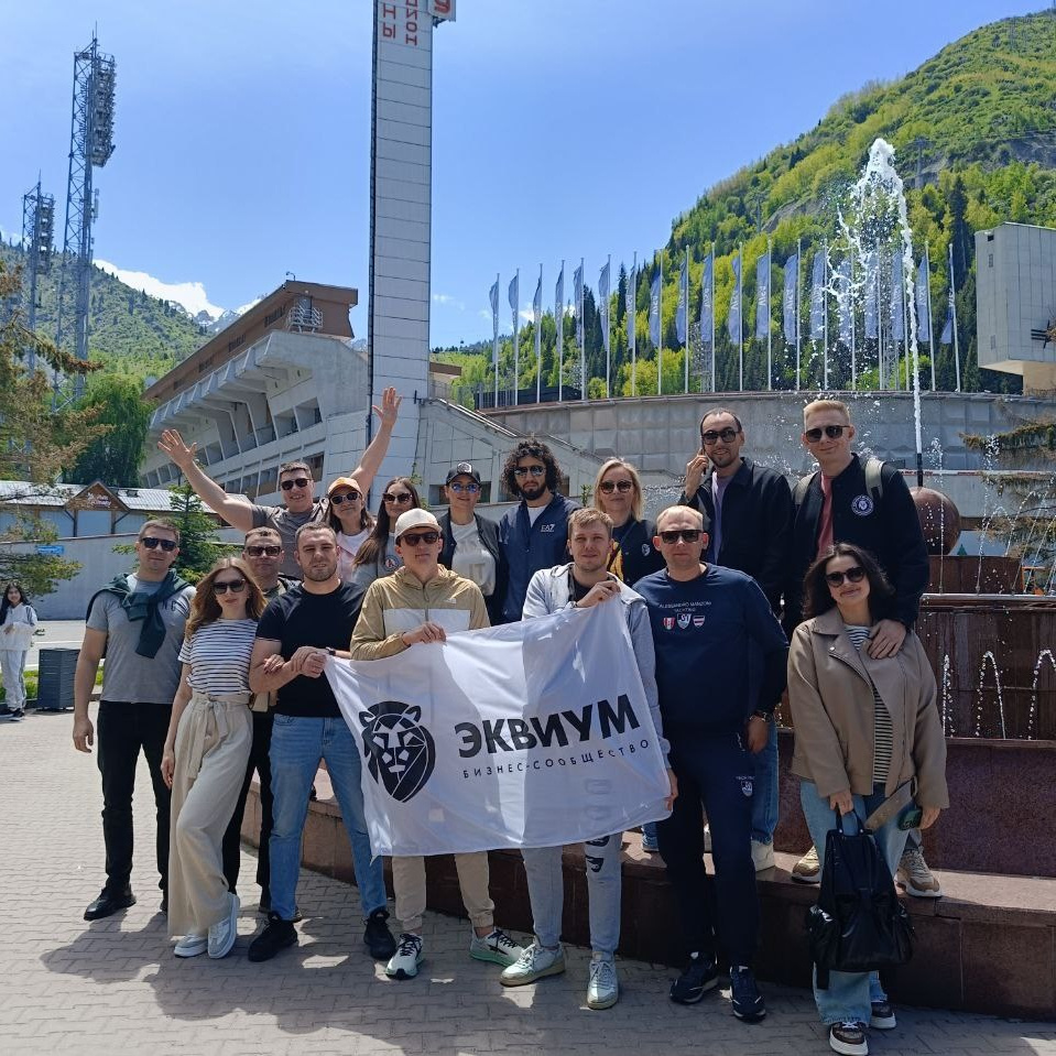 Группа участников корпоративного тура от компании Melon Travel стоит на фоне известного катка "Медеу" в солнечный день. Участники держат белый флаг с надписью "Эквиум. Бизнес-сообщество" и улыбаются. На заднем плане видна архитектура катка, зелёные горы и фонтаны, а также ясное голубое небо, создающее атмосферу веселья и единства.