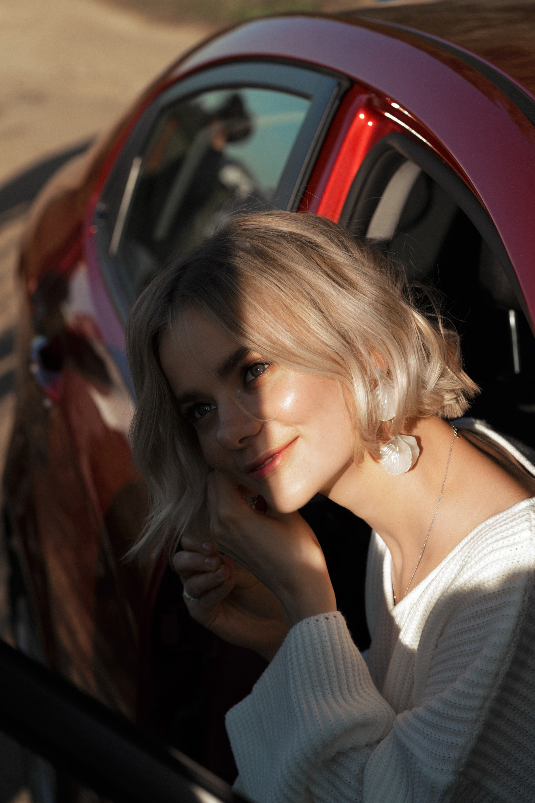 Сидя в красной машине, невеста надевает сережки, жмурясь от яркого солнца.