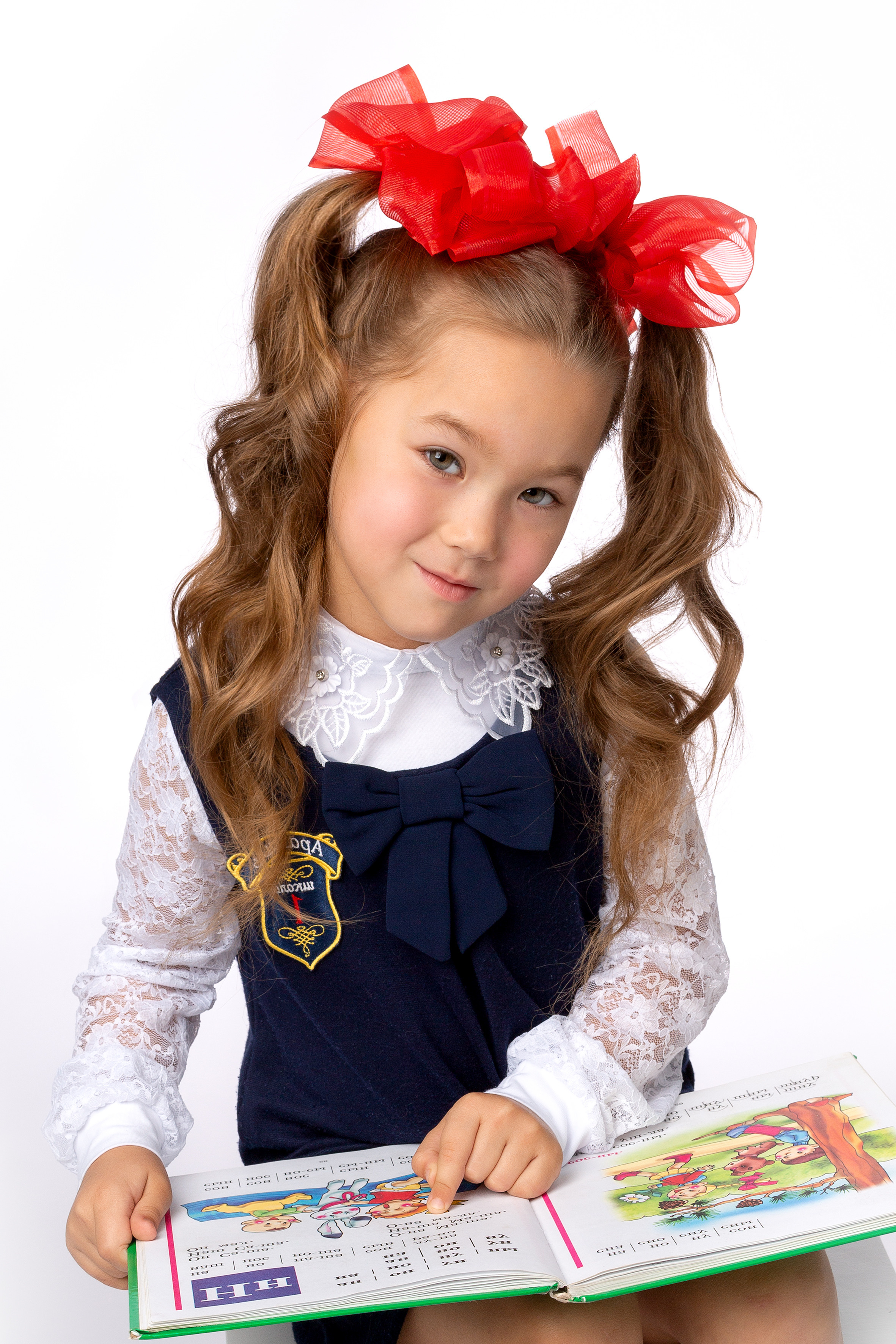 Первоклашка с красными бантами показывает навыки чтения букваря.