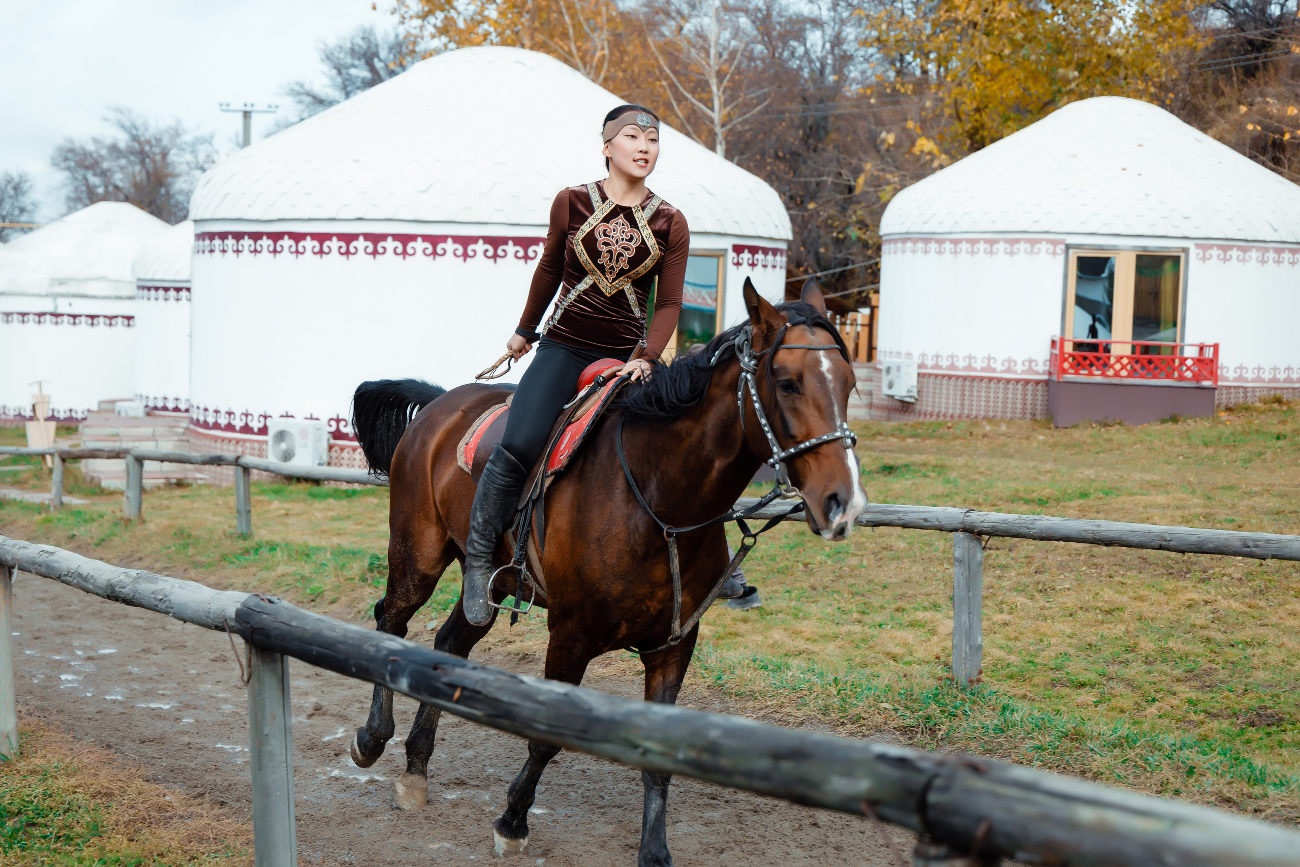 Горное озеро Иссык в Алматинской области, Казахстан, с бирюзовой водой на фоне осеннего леса с яркими золотистыми деревьями, окружённого высокими горами, покрытыми хвойным лесом. На горизонте видны заснеженные вершины под облачным небом