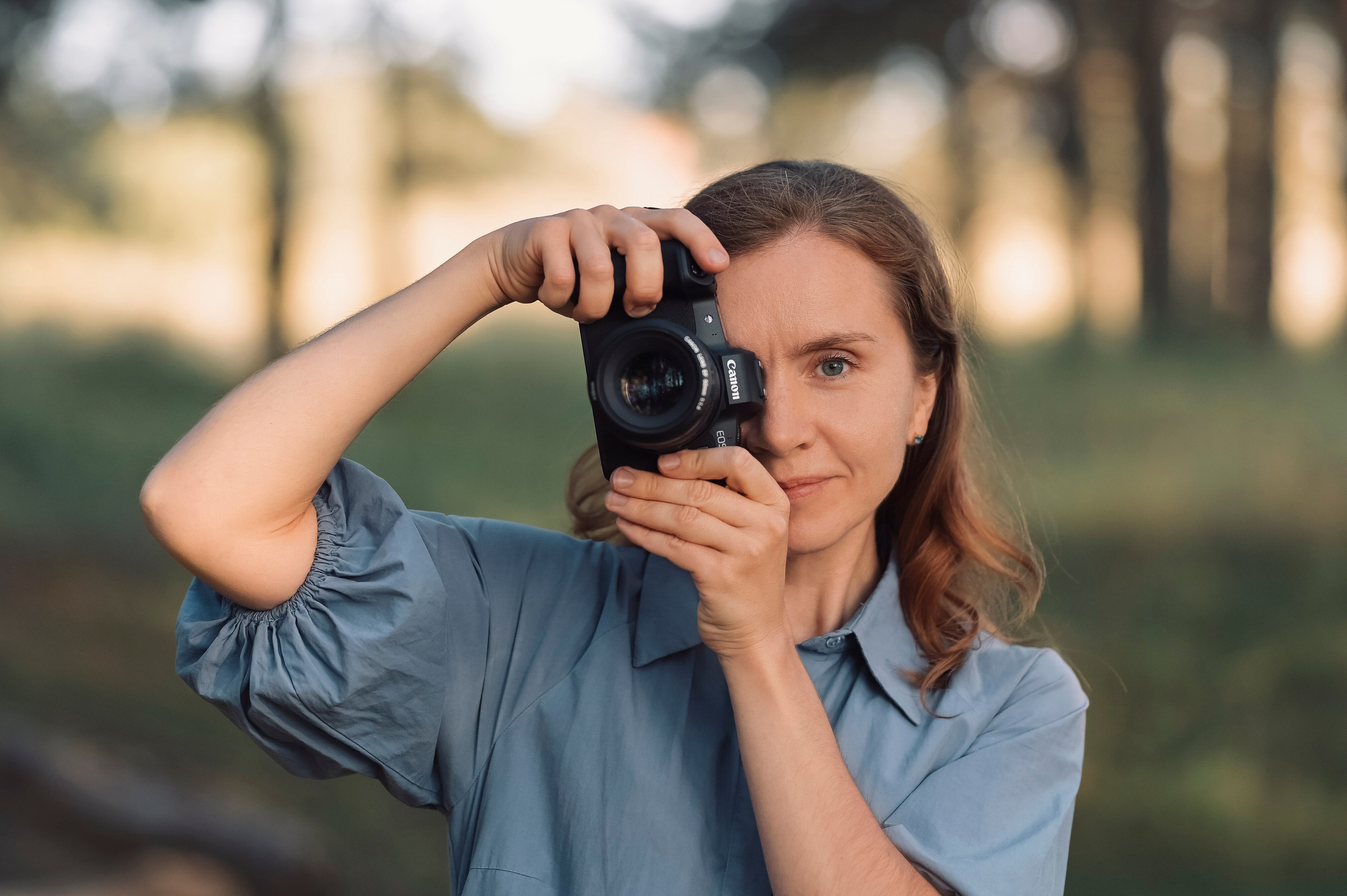 Девушка стоит с фотокамерой в летнем лесу. Камера прикрывает один глаз.
