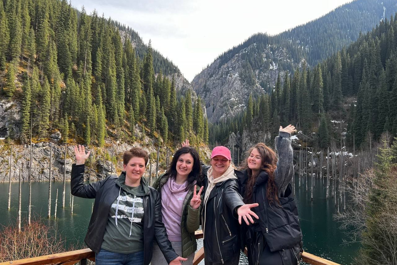 Мужчина и женщина стоят вместе на вершине скалы с видом на пустынный Лунный каньон. Женщина в зелёной футболке, джинсовых шортах и белой кепке, мужчина — в оранжевой футболке, чёрных шортах и кепке. На заднем плане видны скалистые холмы, тёмно-зелёная растительность в долине и драматичное небо с грозовыми облаками, создающее контраст с сухой, песчаной землёй.