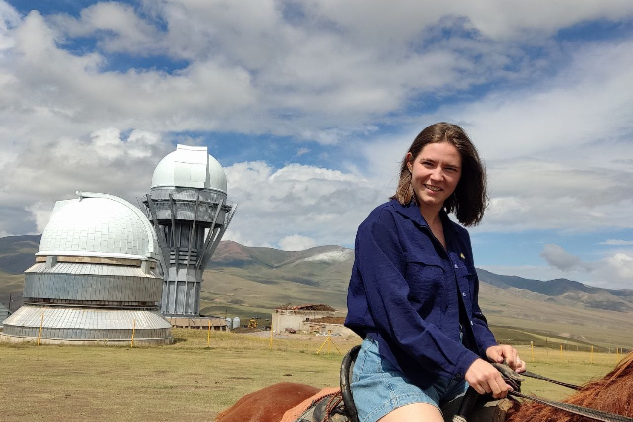 Человек позирует на фоне бирюзового озера Иссык в районе Алматы и гор. Он стоит на большом камне, одет в ярко-зеленую толстовку, черные штаны и светлую обувь, а также оранжевую панаму. В руках держит очки. За ним виднеются деревья, лесистые склоны и горные вершины под облачным небом. Атмосфера спокойная и природная.