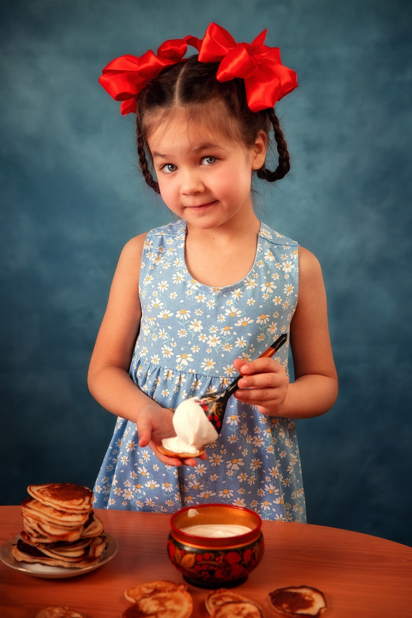 Девочка с красными бантами кушает оладушки со сметаной.