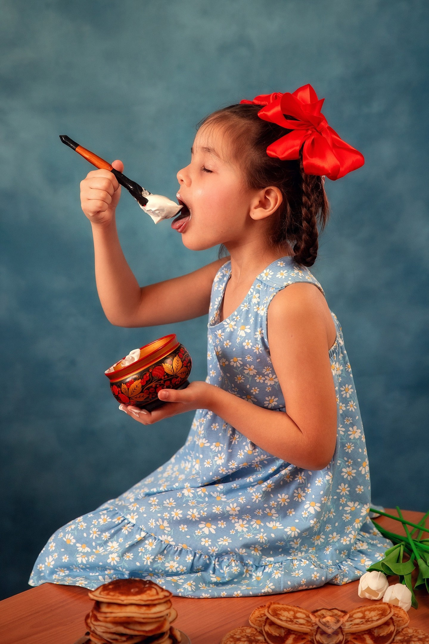 Девочка с красными бантами кушает оладушки со сметаной.