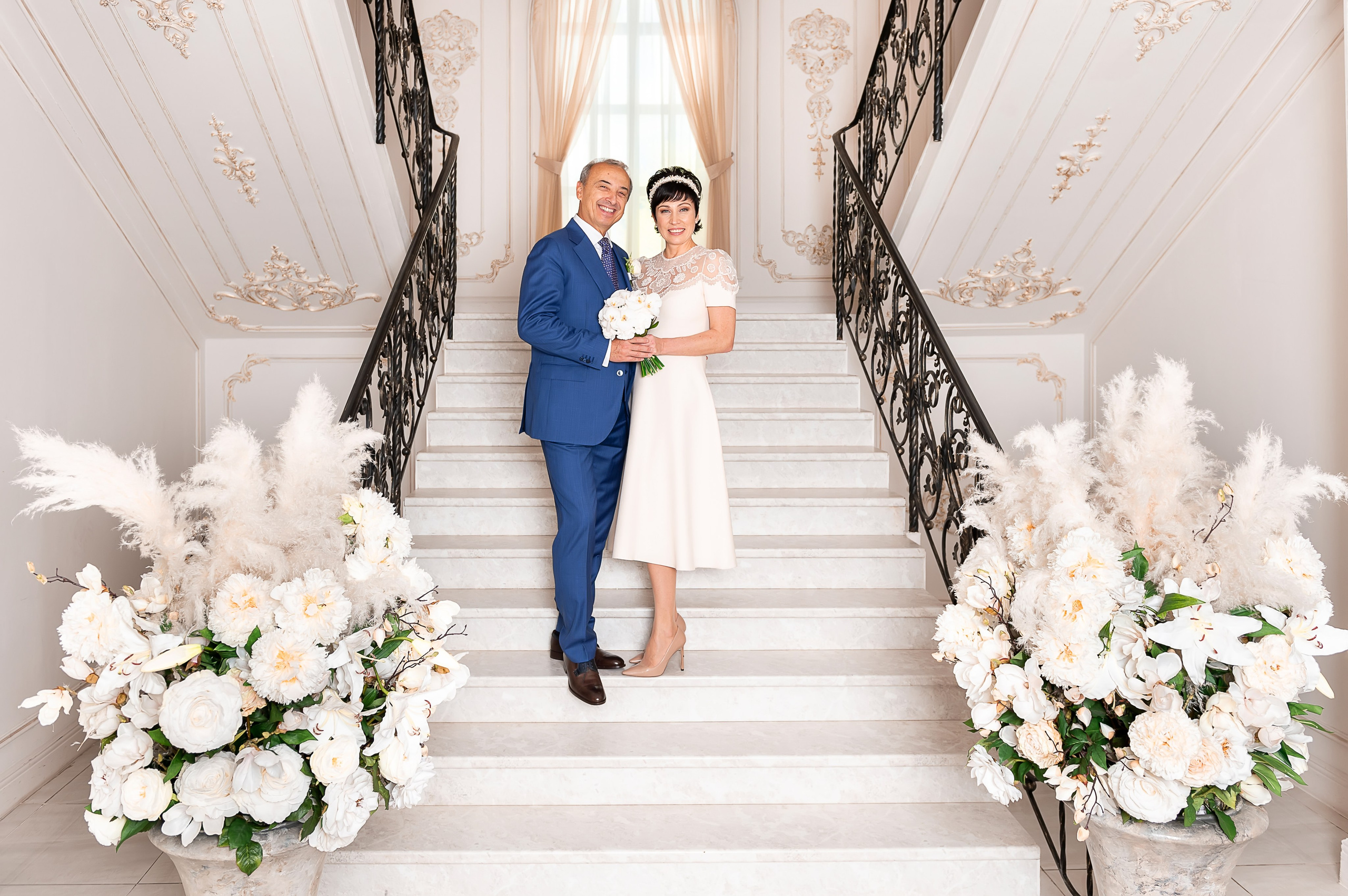 Жених и невеста стоят обнявшись на красивой белой лестнице; справа и слева от них расположены огромные цветочные композиции