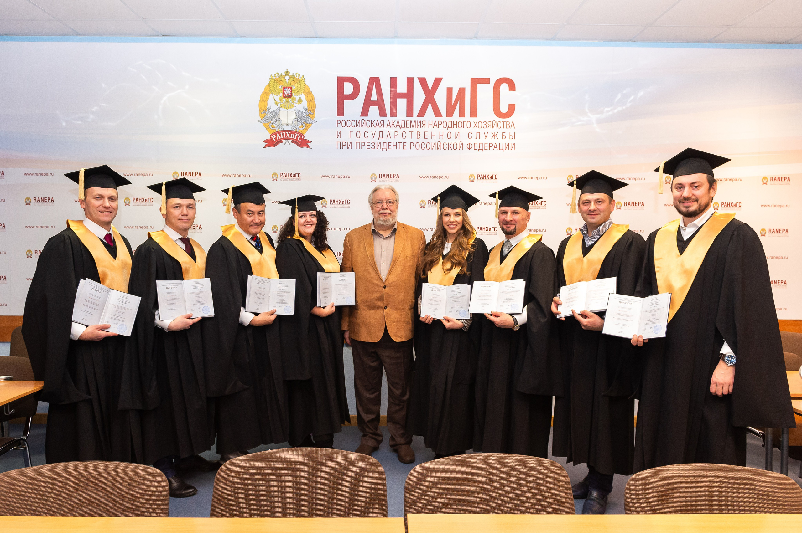 Репортажная фотография выпускников РАНХиГС, которые стоят вместе со своим научным руководителем в аудитории и держат в руках раскрытые дипломы о втором высшем образовании