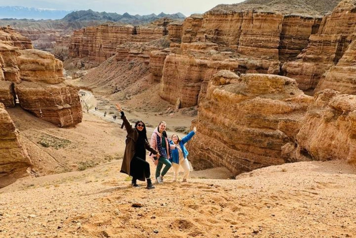 Три человека позируют на склоне холма с видом на Долину Замков Чарынского каньона. Они стоят на фоне крупных скальных образований, характерных для пустынного ландшафта, с глубокими ущельями. Дорога внизу петляет между высокими скалами. Горы виднеются вдали на горизонте под светлым небом. Люди улыбаются и радостно поднимают руки вверх.