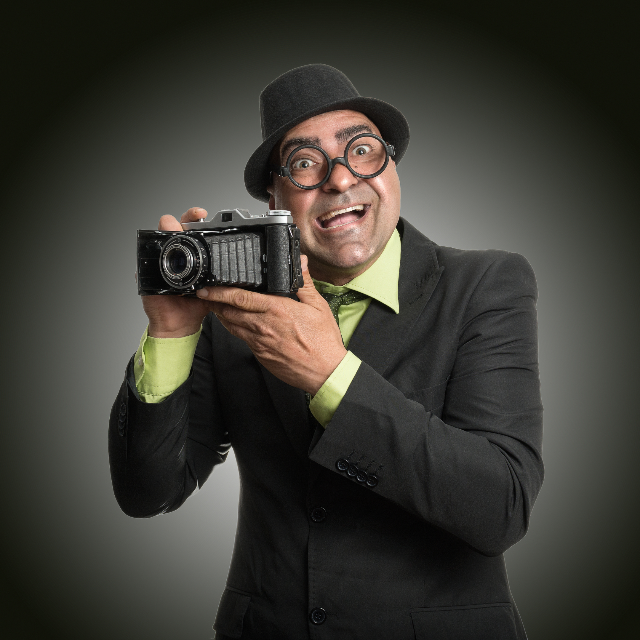 Мужчина в шляпе и очках с широкой улыбкой держит старую плёночную камеру, позируя перед объективом