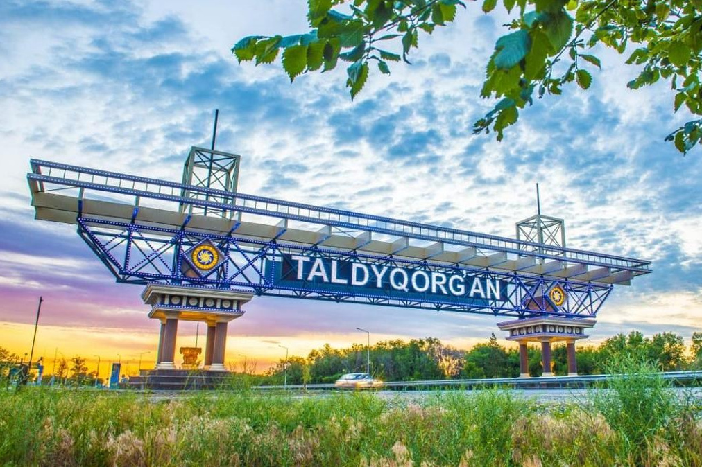 Величественный Чарынский каньон в районе Алматы с ярко-красными скалами, пересечённый извилистой дорогой, на которую падают длинные вечерние тени. Захватывающее сочетание красочных утёсов, зелёных склонов и драматичного неба создаёт эффектный пейзаж