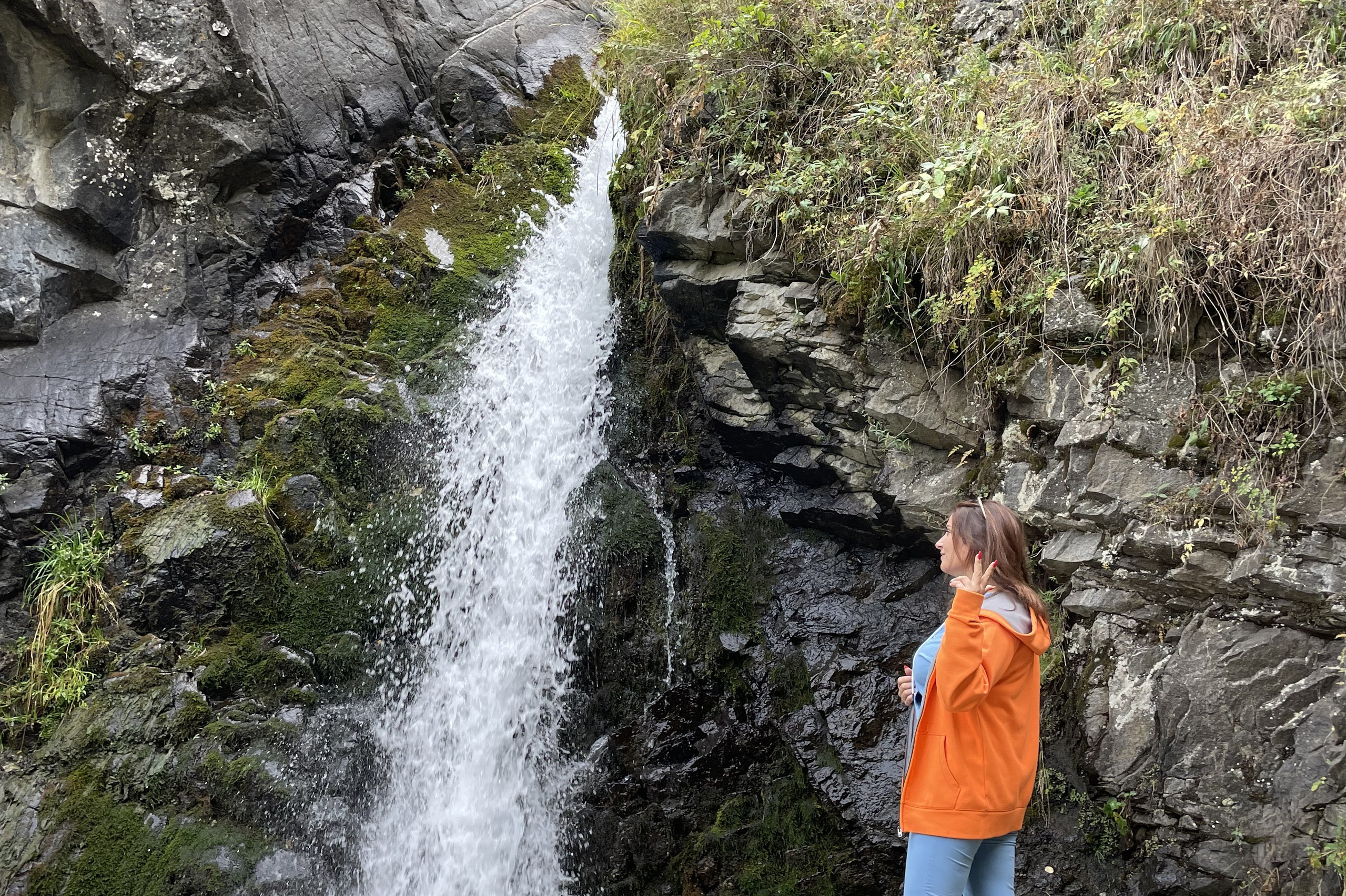 Пейзаж, напоминающий лунный, с извилистой рекой, окруженной густой зеленью и деревьями на фоне пустынных скалистых гор Лунного каньона. Солнечный день с ярко-голубым небом, украшенным небольшими облаками, подчеркивает природную красоту местности. Река Чарын образует небольшие островки, покрытые травой и кустарником.