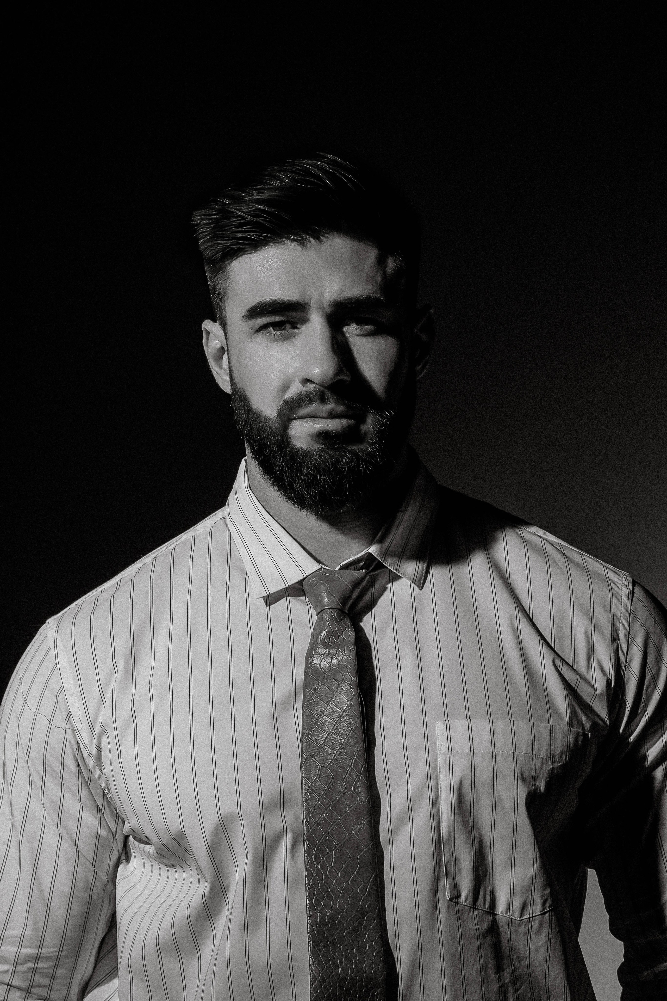 Парень в белой полосатой рубашке с узким галстуком со стильной прической и выглаженной бородой на темном фоне в зале с циклорамой