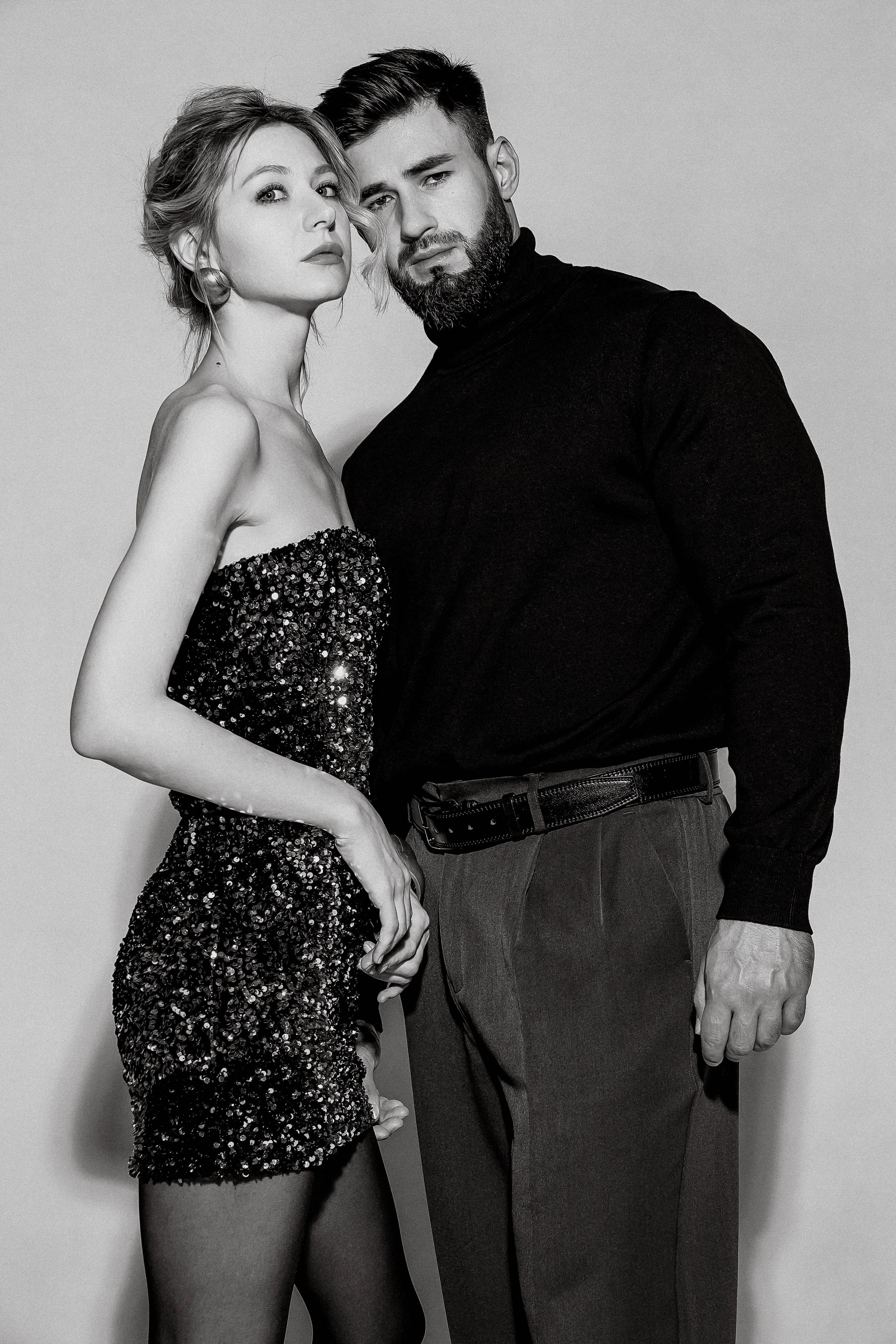 Парень со стильной прической и бородой в черной водолазке прижимает к себе девушку в блестящем коротком платье - фото в ч/б