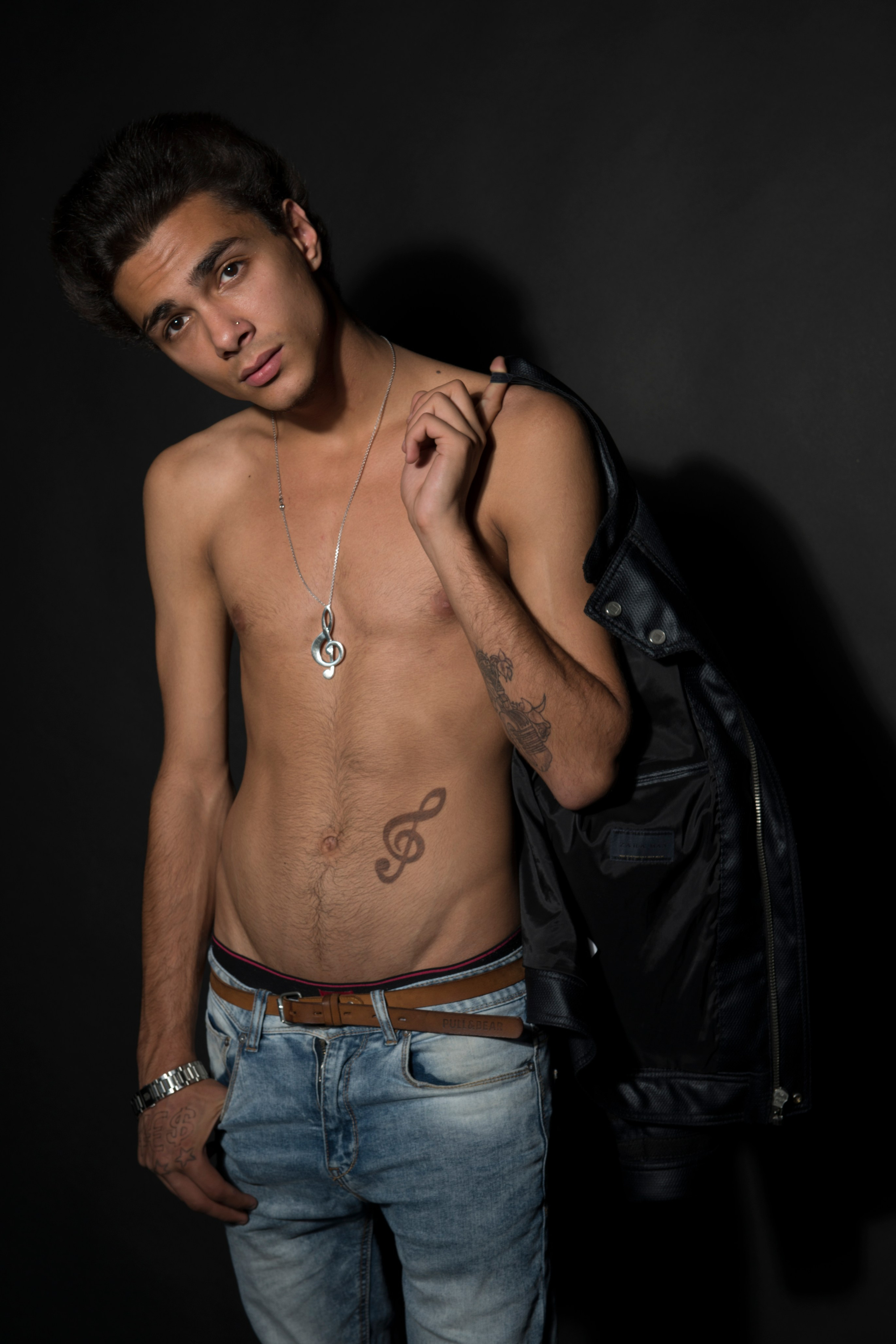Полуобнажённый мужчина стоит с курткой на плече и татуировкой на животе.