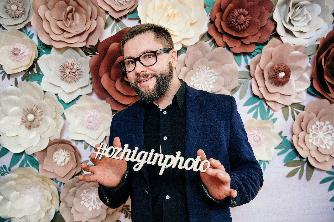 мужчина в очках на фоне цветов в черной рубашке и пиджаке улыбается и держит хэштеги ozhiginphoto , на лице у мужчины борода. Фотограф Денис Ожигин Краснодар, пос.&nbsp;Южный (Динской район) с&nbsp;выездом по&nbsp;Краснодару и&nbsp;Краснодарскому краю