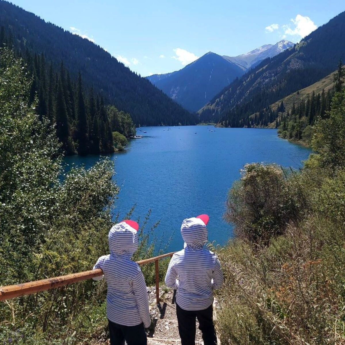 Два ребёнка из Алматы в одинаковых полосатых толстовках с красными козырьками стоят на лестнице, ведущей к живописному озеру Кольсай в Казахстане. Озеро окружено горами, покрытыми густым лесом. Ясное голубое небо подчёркивает яркий цвет воды и зелень природы вокруг. Вид открывается на тихий и спокойный пейзаж с вершиной горы вдали.
