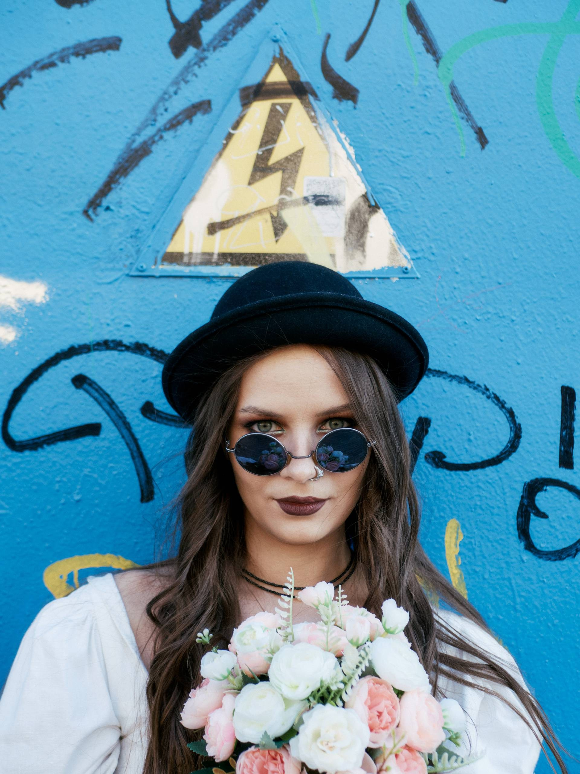 Девушка в чёрной шляпе и солнцезащитных очках держит букет цветов на фоне яркой стены с граффити и знаком молнии