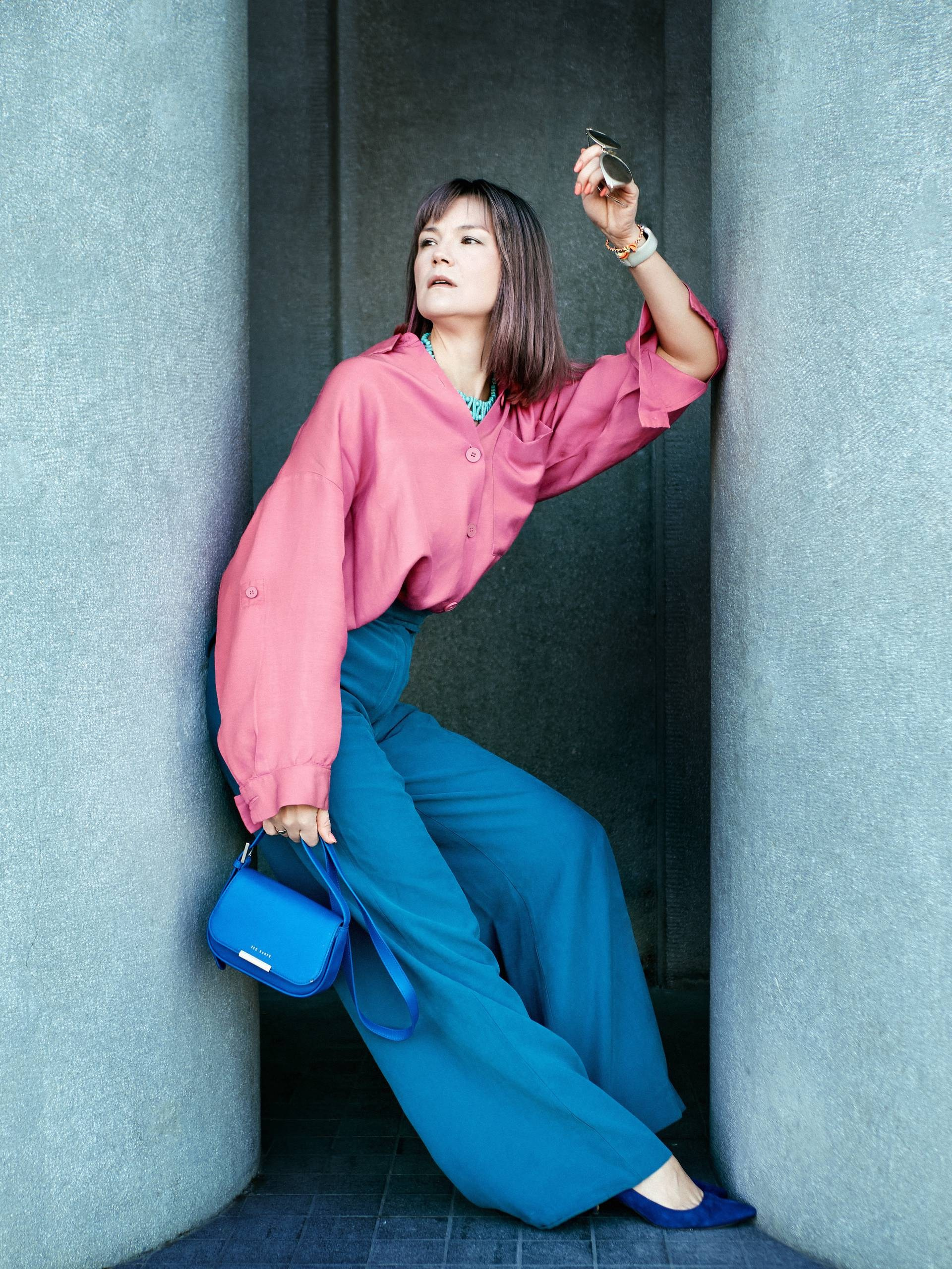 Девушка в ярко-розовой рубашке и широких синих брюках позирует между бетонными колоннами с синей сумкой в руке