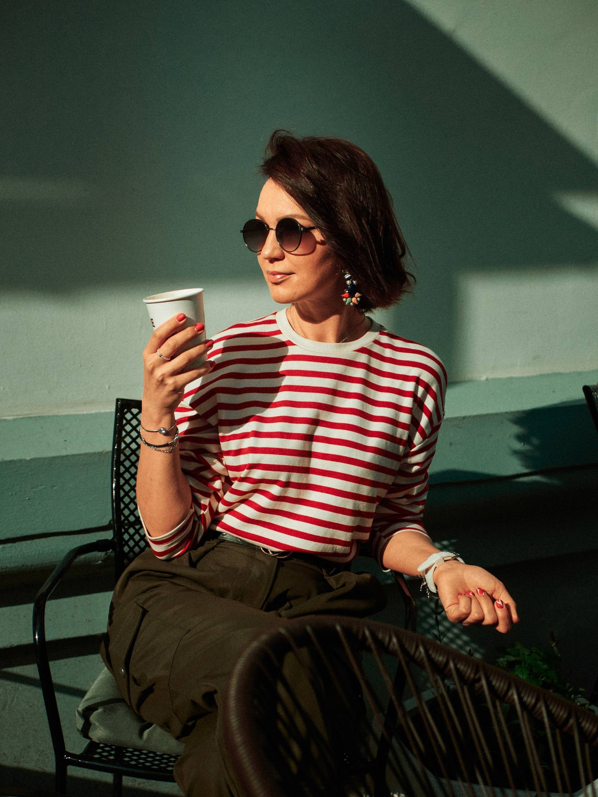 Стильная девушка в полосатой футболке и солнцезащитных очках сидит на улице с чашкой кофе в руке, в лучах тёплого света