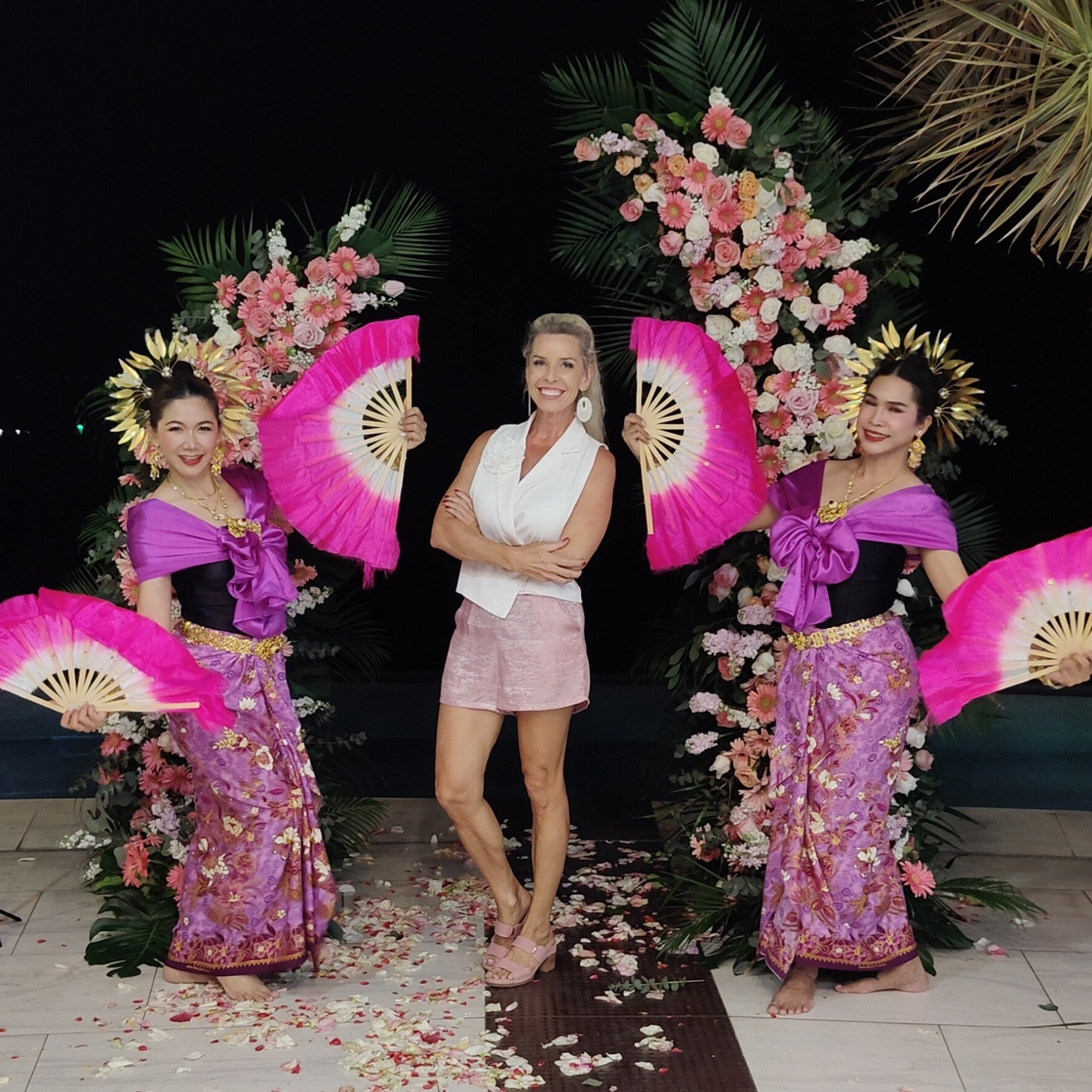 Traditional Thai dancers in colorful pink costumes performing with decorative fans for a female guest during a tropical wedding celebration in Koh Samui, Thailand.
