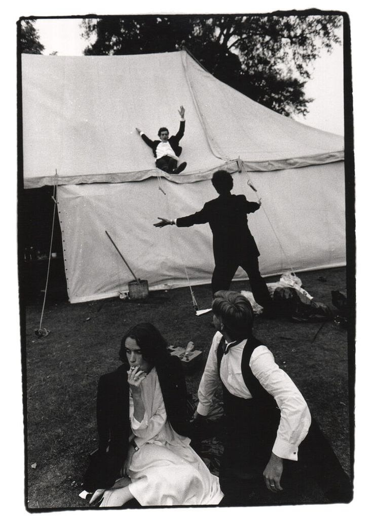 Черно-белое фото свадьбы, пара сидит на переднем плане, двое парней на фоне развлекаюся и прыгают на шатёр