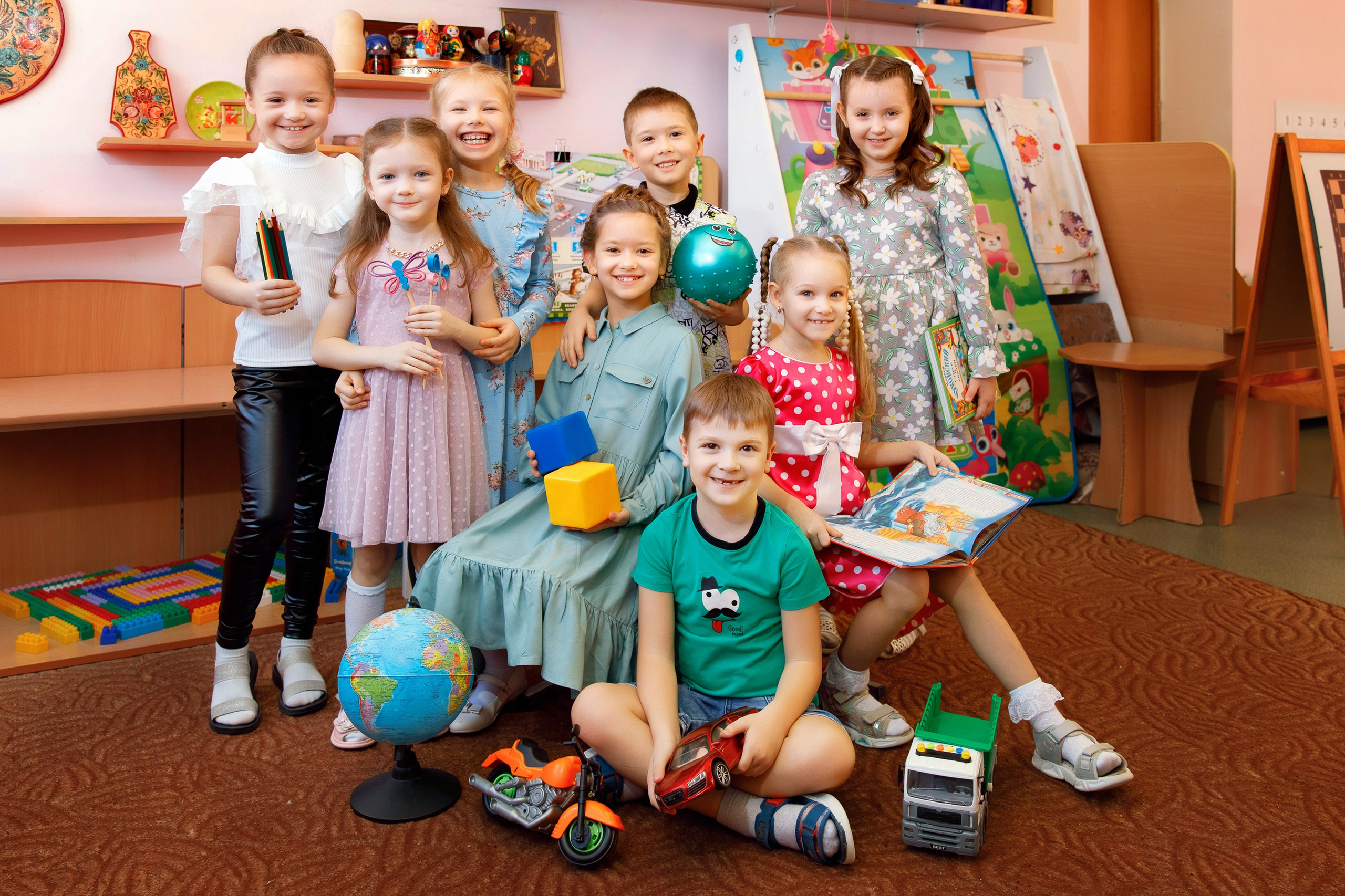 Группа детей улыбается, стоя в группе с игрушками и глобусом.  Дети позируют с улыбками в детском саду, держа книги и игрушки. Веселые дети в яркой одежде сидят среди игрушек и книг. Дети держа в руках различные предметы.  Дети демонстрируют учебные принадлежности и игрушки.