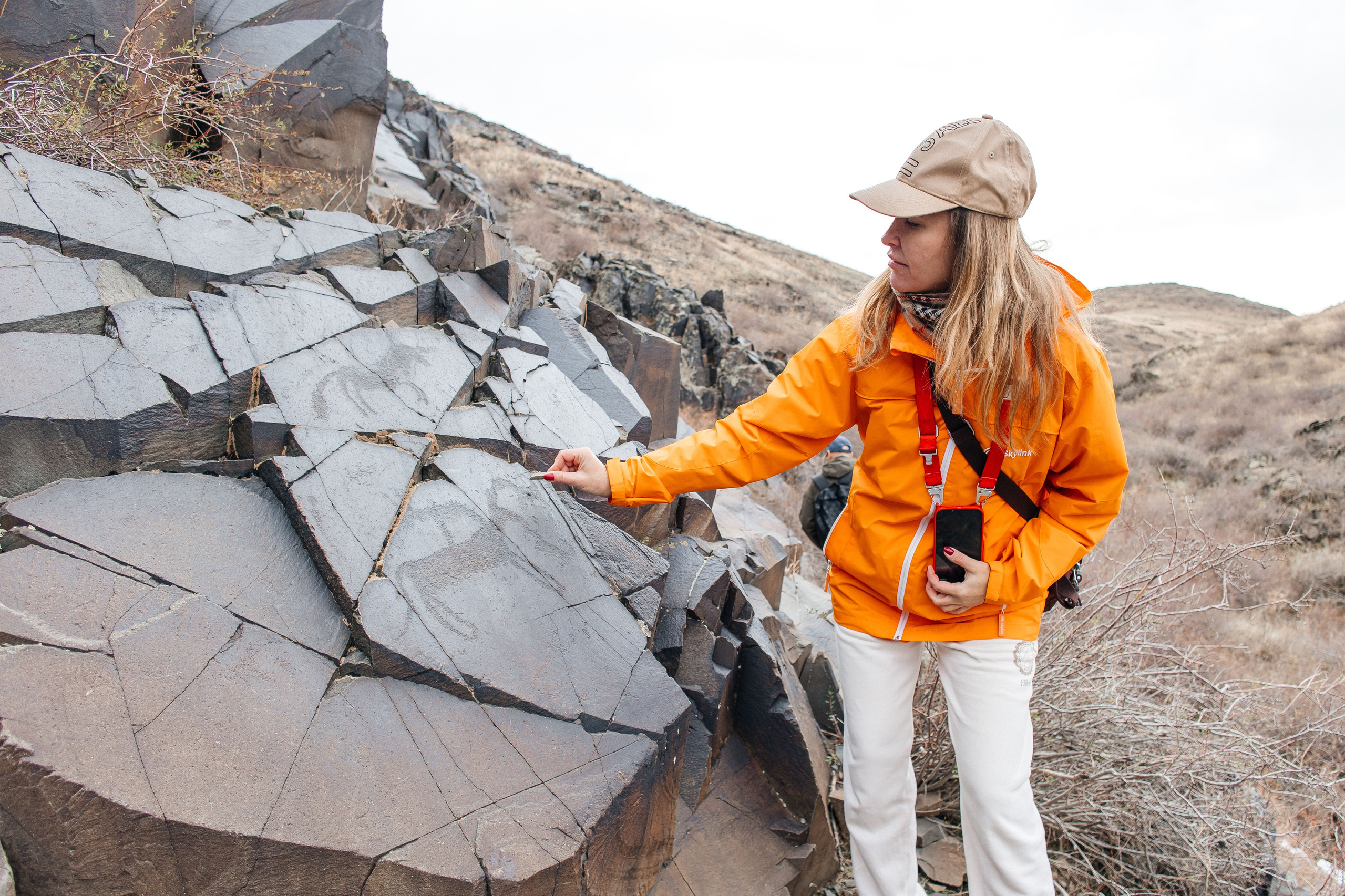 Человек позирует на фоне бирюзового озера Иссык в районе Алматы и гор. Он стоит на большом камне, одет в ярко-зеленую толстовку, черные штаны и светлую обувь, а также оранжевую панаму. В руках держит очки. За ним виднеются деревья, лесистые склоны и горные вершины под облачным небом. Атмосфера спокойная и природная.