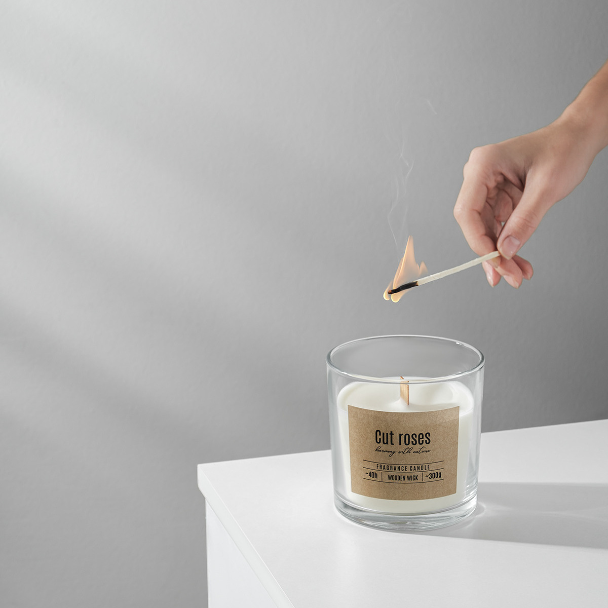 Атмосферная фотография для бренда, продающего свечи, изделие в стеклянной емкости в лучах света, фитиль поджигает рука с длинной спичкой.