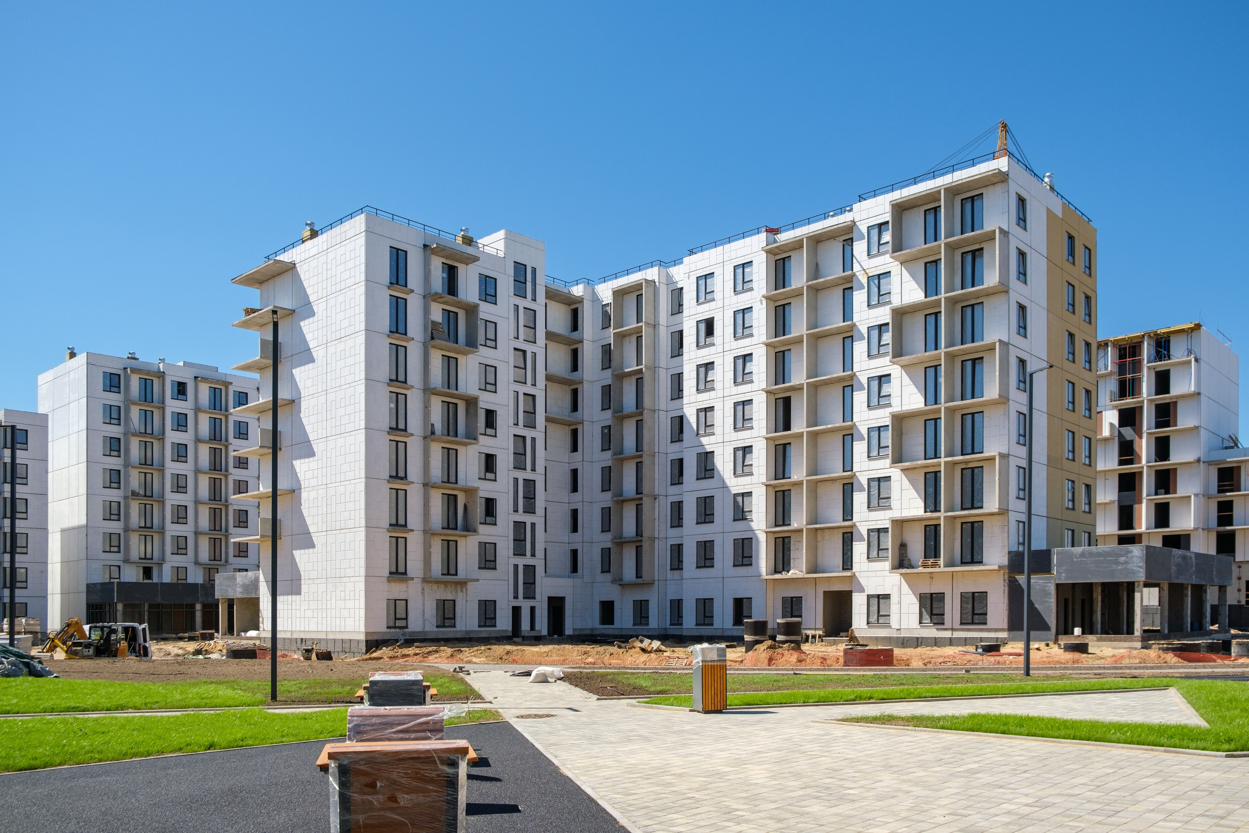 На изображении представлен современный жилой комплекс, состоящий из нескольких многоэтажных зданий с белыми и серыми фасадами. Комплекс окружён открытыми пространствами, где установлены уличные фонари и есть тротуары для пешеходов.