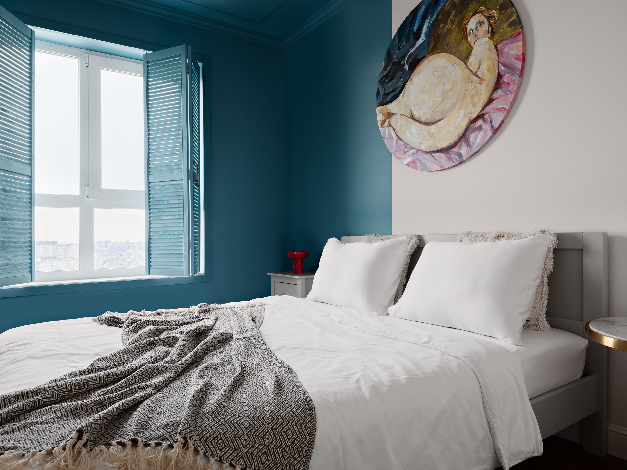 Интерьер спальни с большими окнами и голубыми ставнями, белой кроватью, мягким текстильным покрывалом и картиной над изголовьем. Дизайнер Антон Логинов, стилист Алёна Мощенская и фотограф Сергей Денисенко подчеркнули современную эстетику.