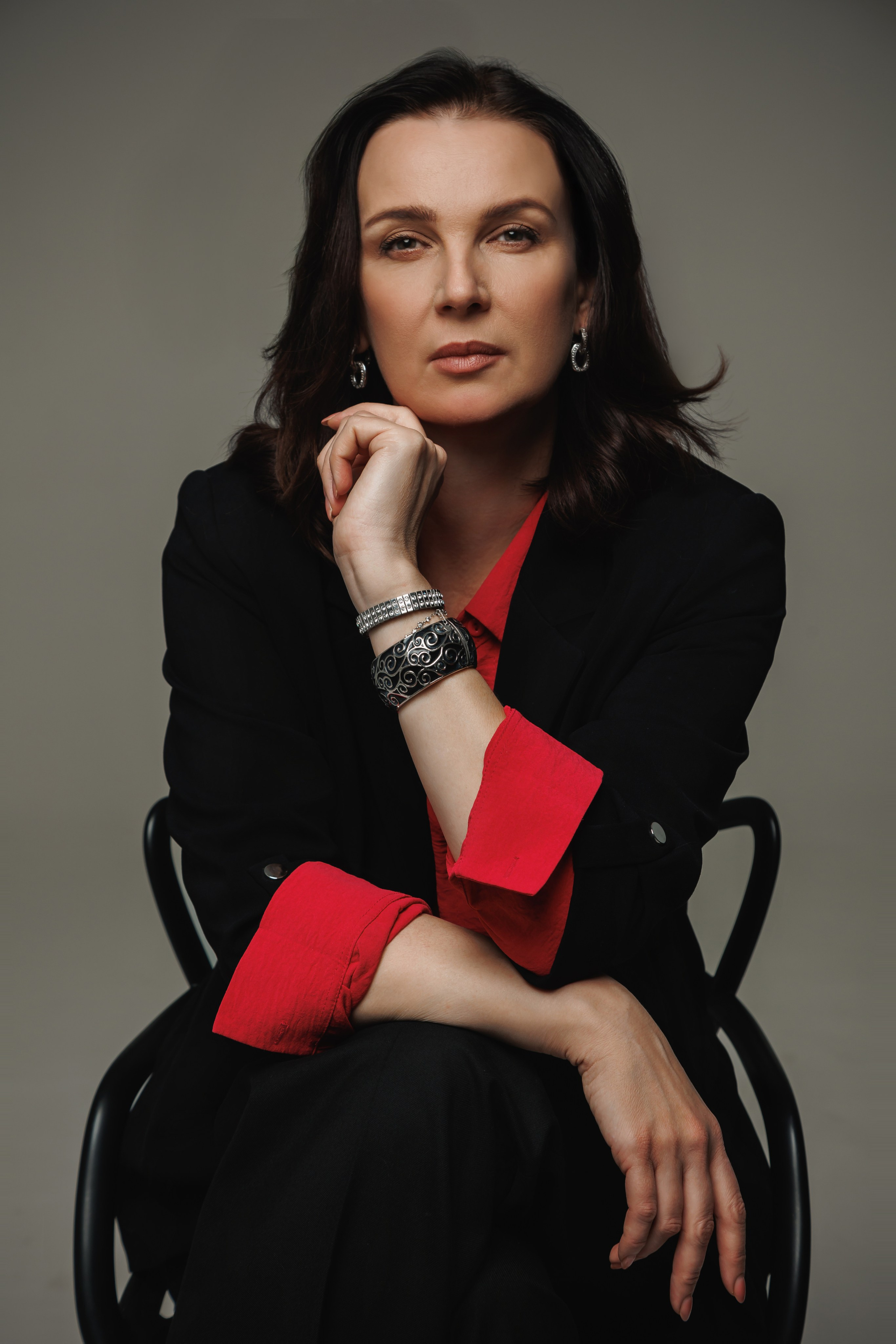 Прямой контактный портрет: женщина-психолог сидит на стуле и смотрит прямо в камеру на сером фоне.