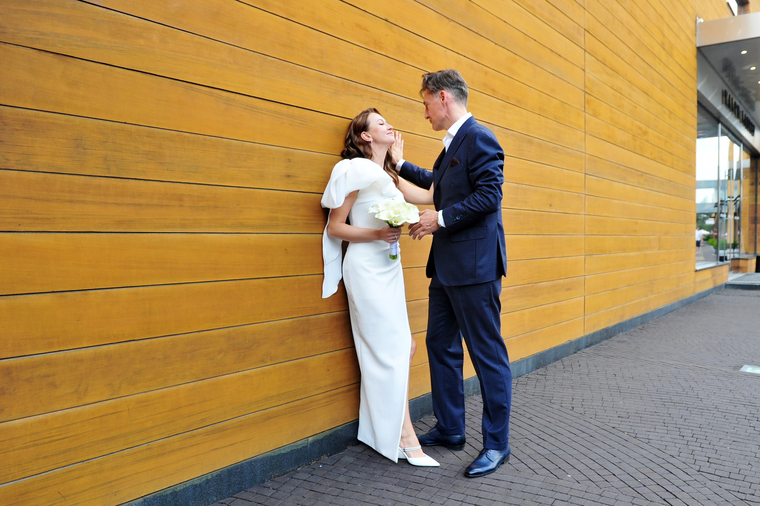 Свадьба в ЗАГСе Барвиха Luxury Village летом, жених и невеста стоят у деревянной стены здания