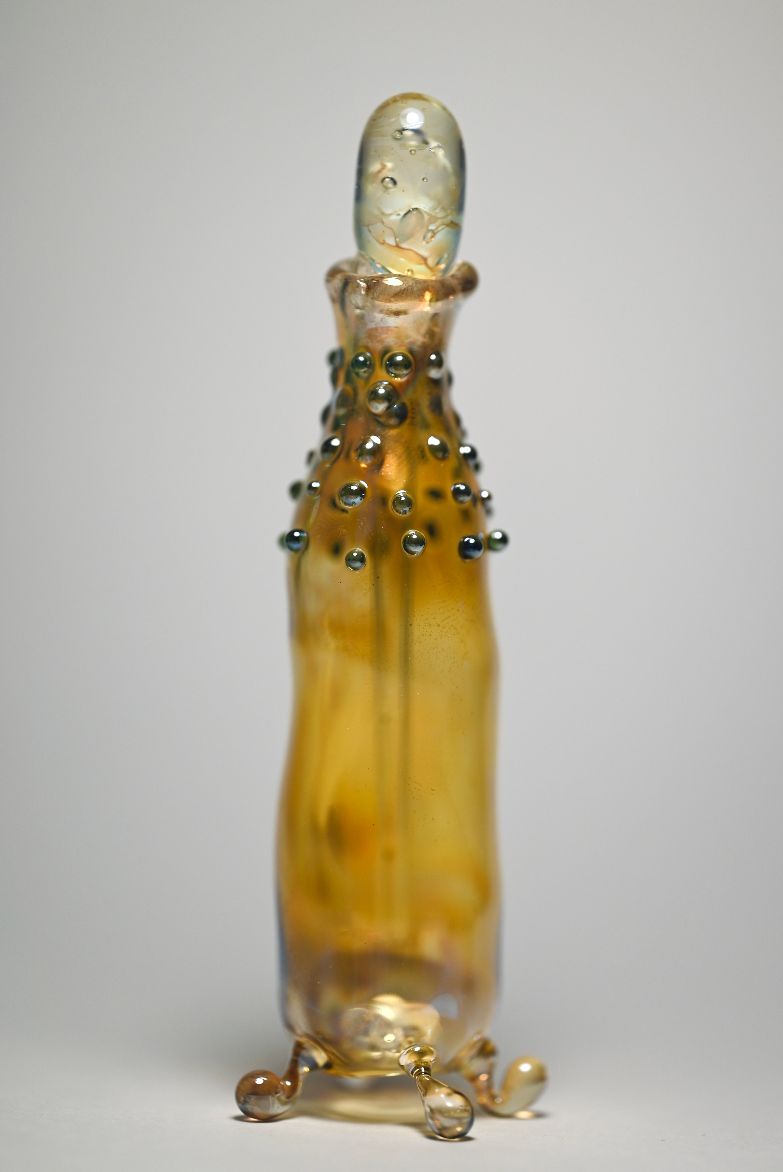 alt="Стеклянная скульптура в виде вытянутой бутылки с золотистым оттенком и округлыми выступами по всей поверхности."