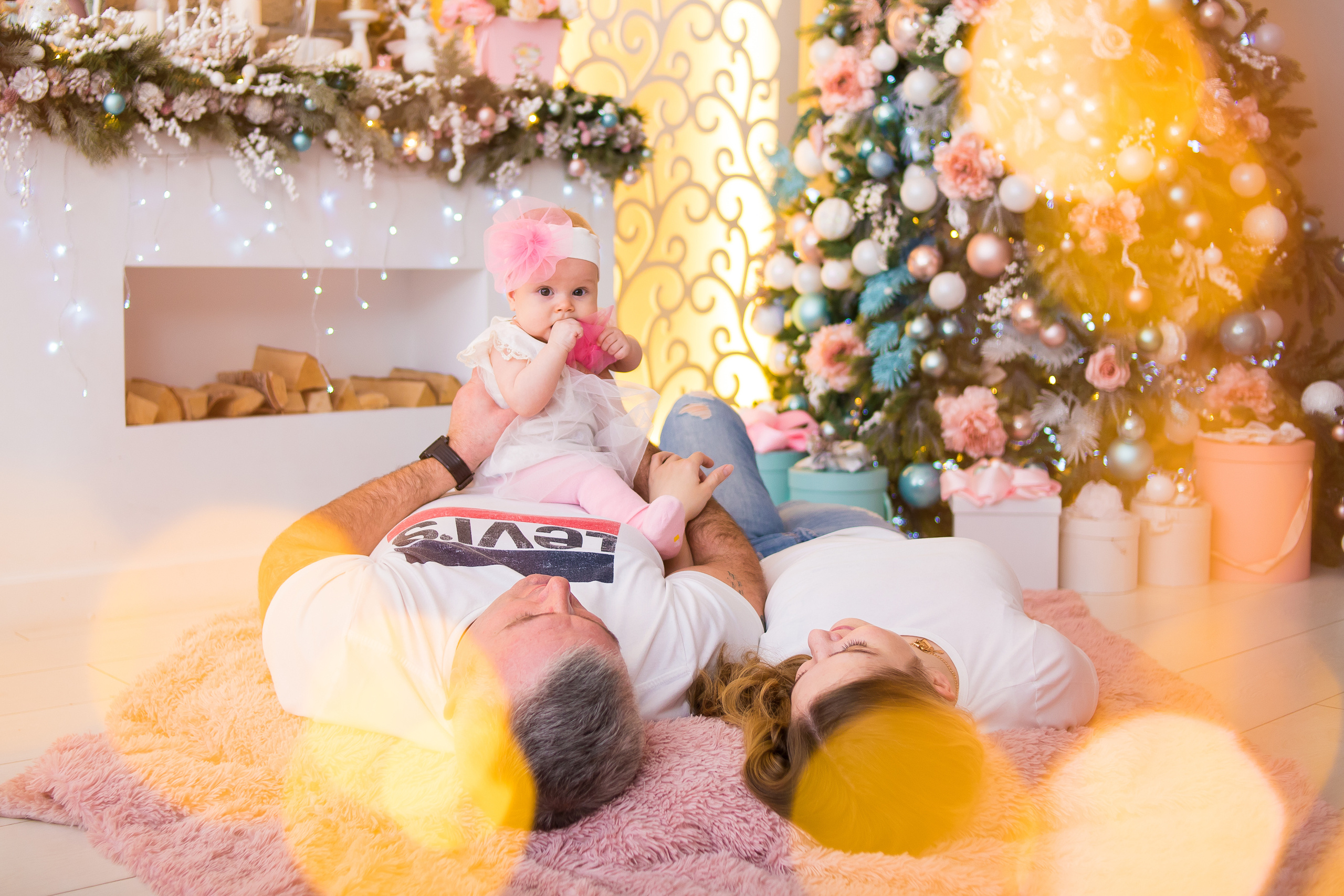 Новогодняя семейная фотосессия с грудничков, на переднем плане размытые огоньки, сзади камин и новогодняя ёлка. 