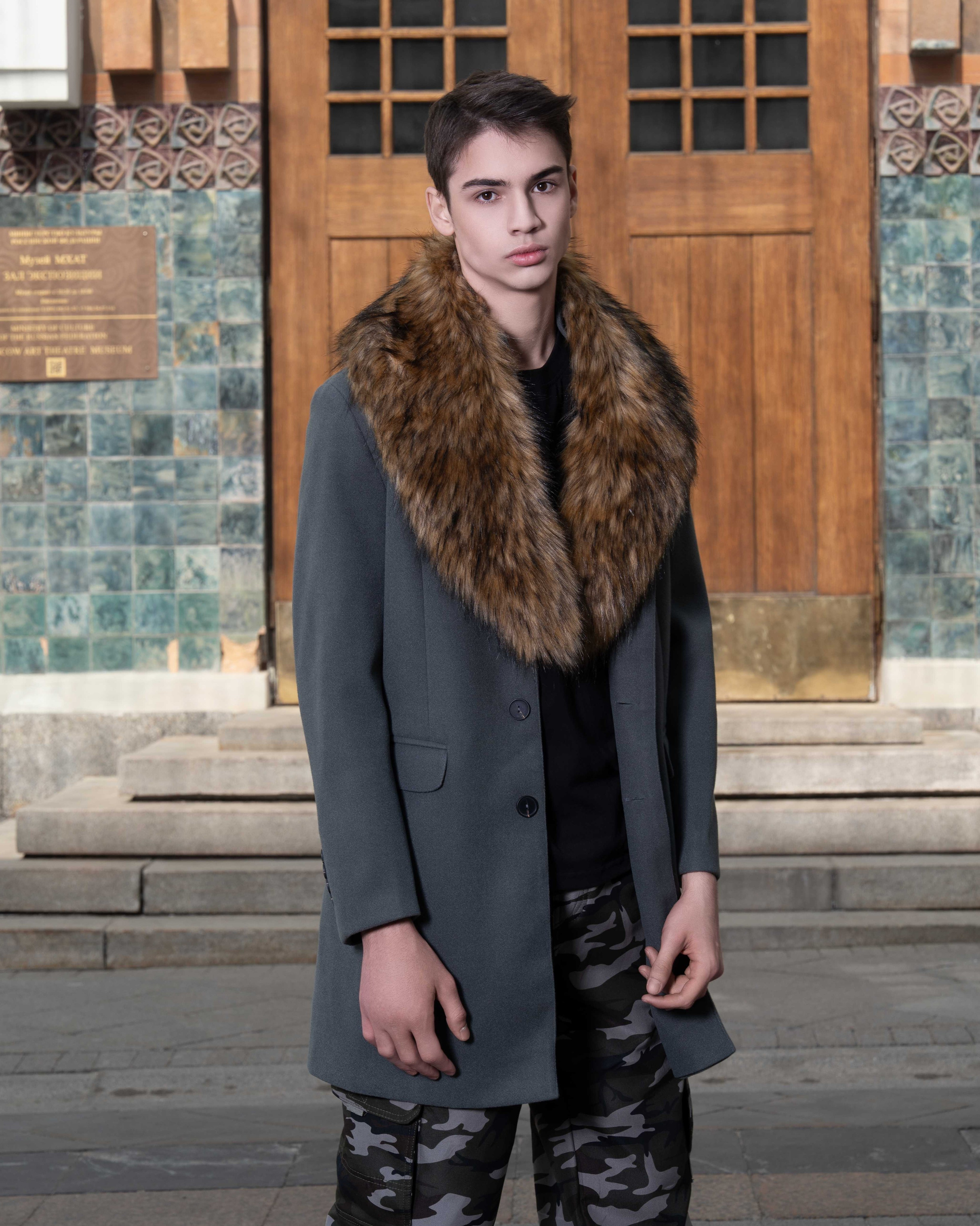 На фотографии изображен молодой человек, который стоит на фоне деревянной двери и каменной стены с мозаикой. Он одет в серое пальто с меховым воротником, который придает образу элегантность и тепло. Под пальто видна черная футболка, а на ногах у него камуфляжные брюки, что добавляет образу немного непринужденности и стиля. Молодой человек смотрит прямо в камеру, его взгляд серьезен и сосредоточен. Освещение на фотографии мягкое, что подчеркивает детали его одежды и создает приятную атмосферу.