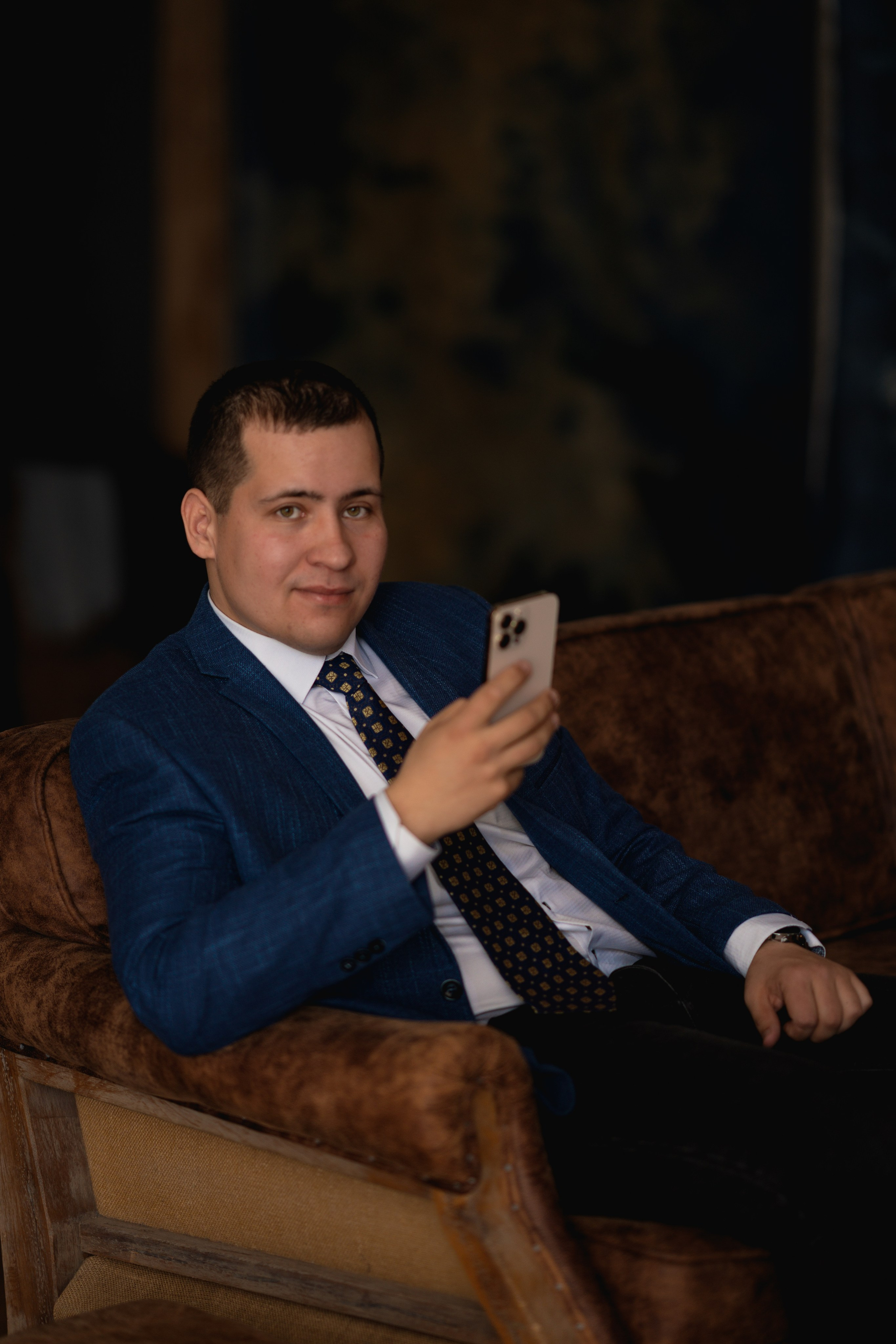 Мужчина в строгом костюме с телефоном в руках сидит на коричневом кожаном диване