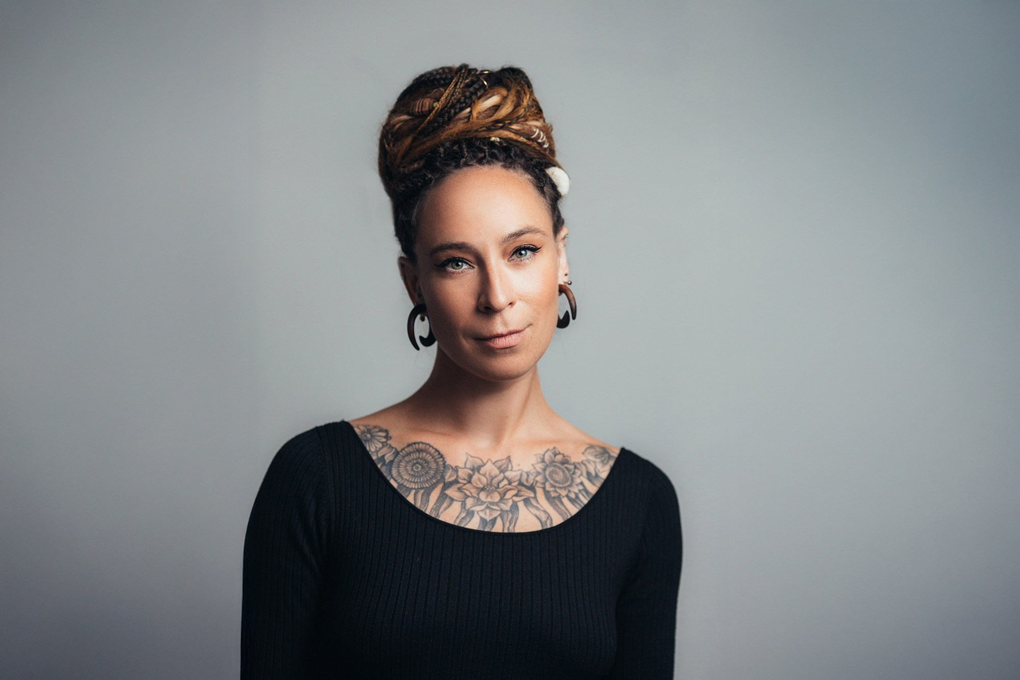 Портрет женщины с дредами, собранными в высокую прическу, и крупными серьгами-растяжками. На ключицах видна детальная татуировка с цветочными и геометрическими мотивами. Спокойный взгляд направлен в камеру, фон нейтральный серый. Контент фотограф Галина Скорик.