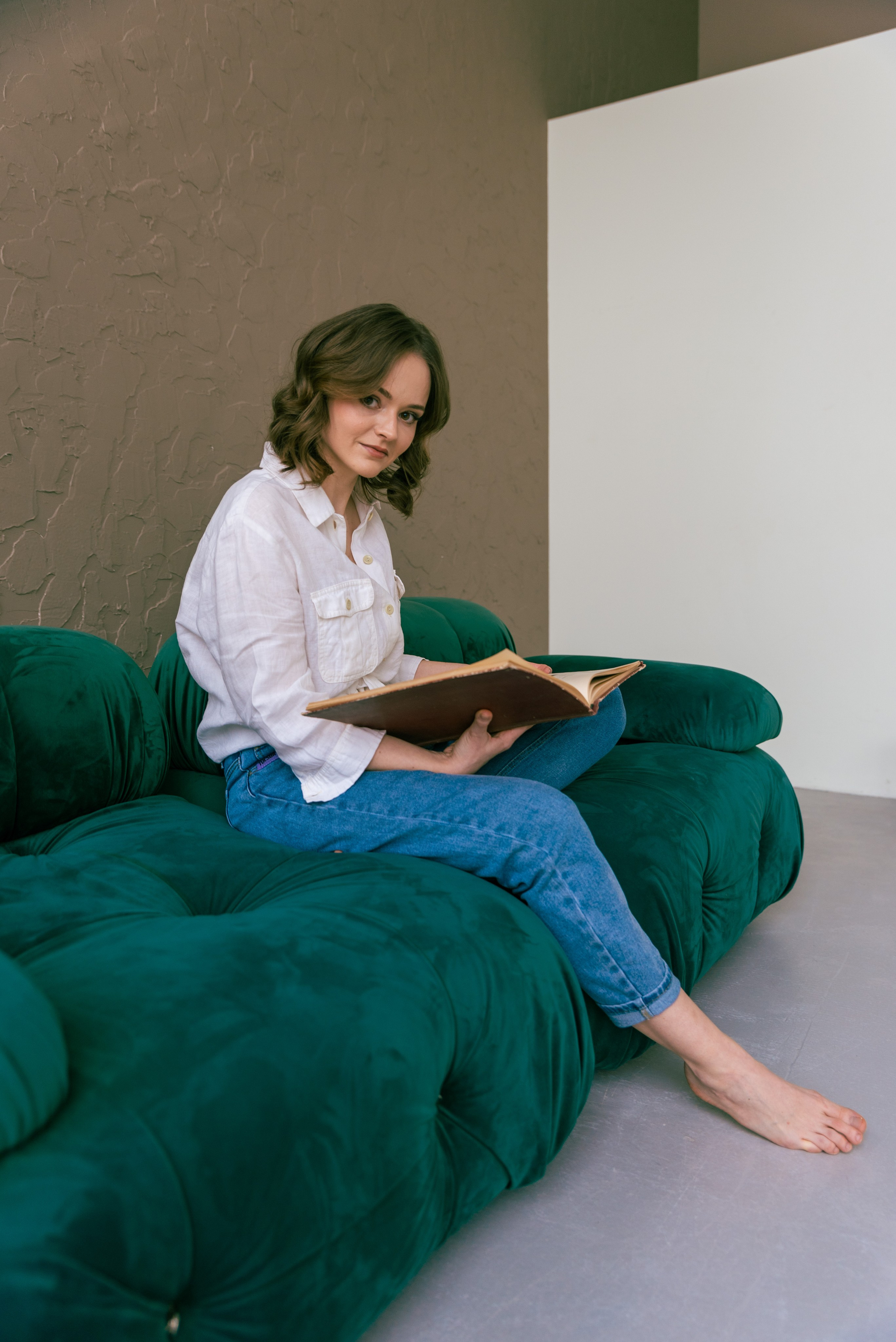 Фото в интерьере. Девушка в белой рубашке и джинсах сидит на диване, держит в руках книгу.