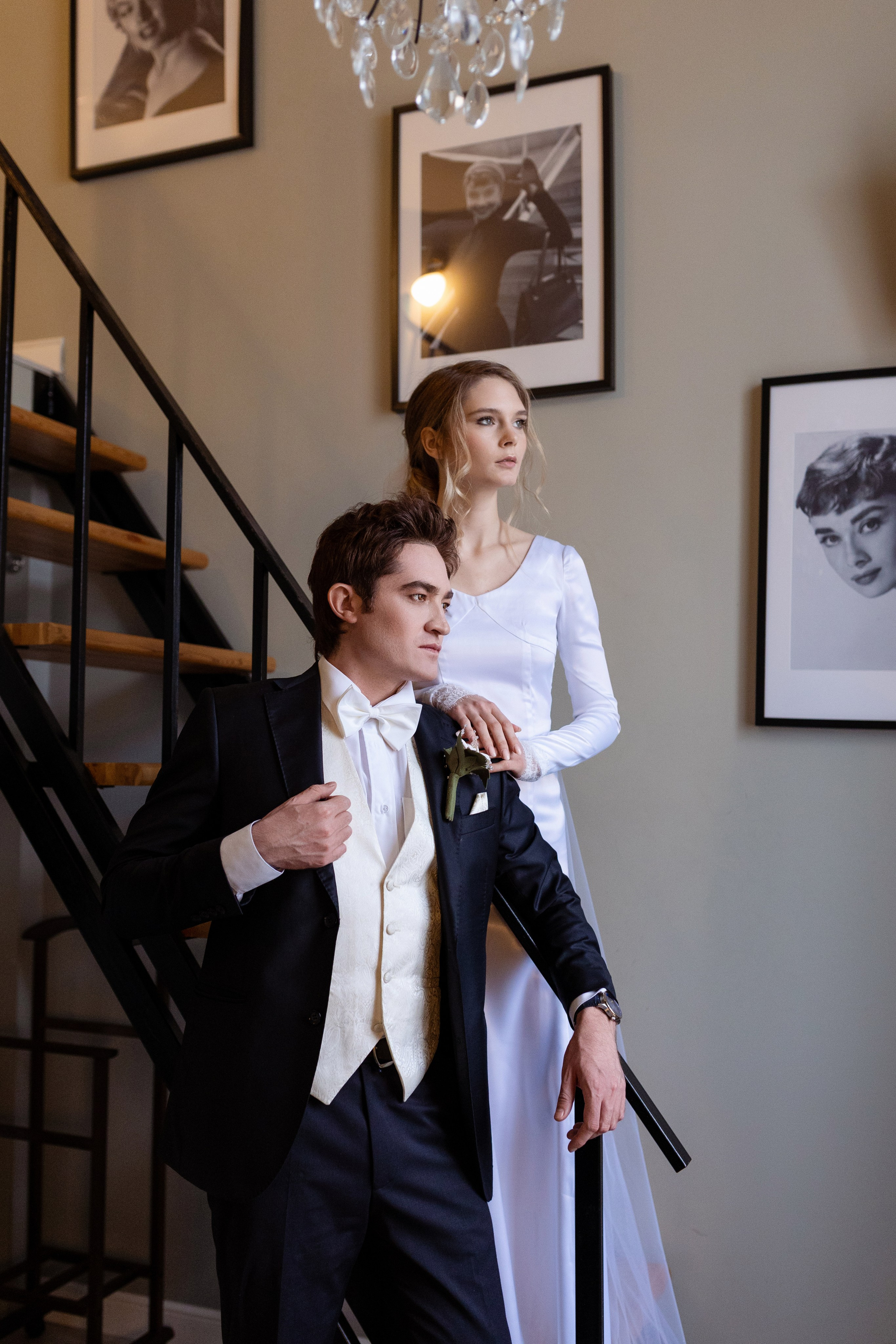 Невеста стоит на лестнице сзади жениха На фоне фотографий развешанных на стене.