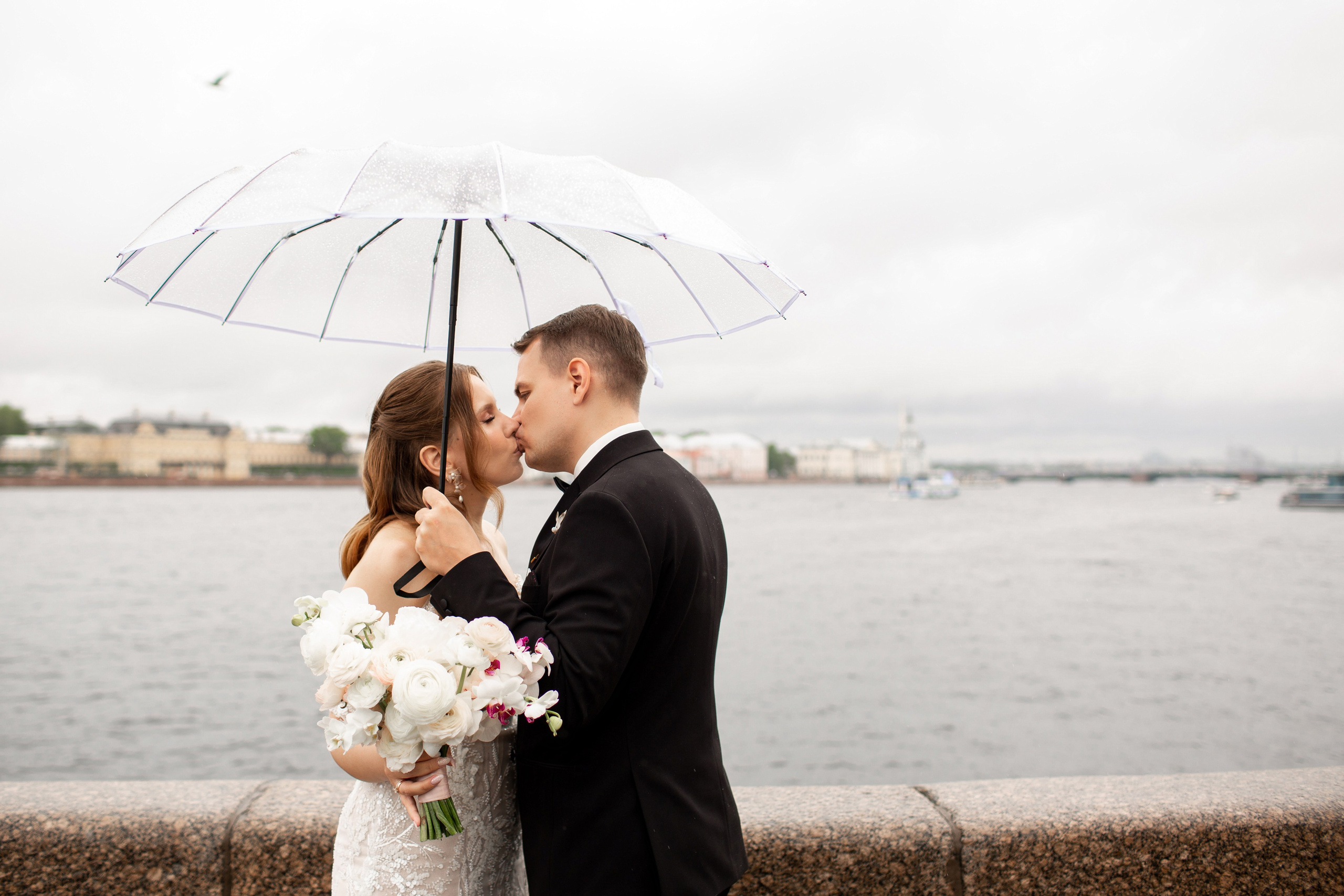 Классическая петербургская фотография: красивая пара в свадебных нарядах целуется на набережной под зонтом.