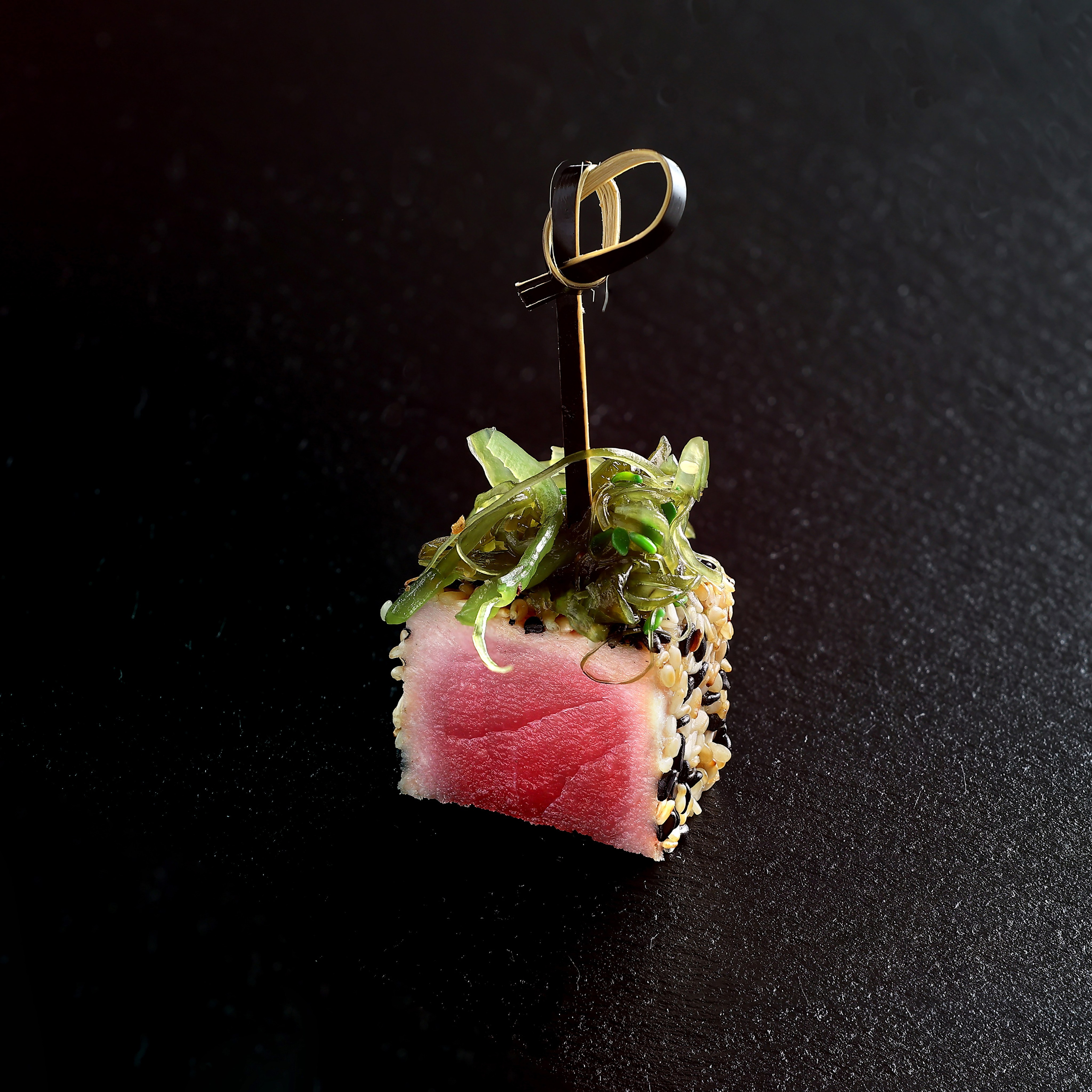 Запечённый медиум тунец в кунжуте с морской капустой на шпажке, на тёмном фоне.