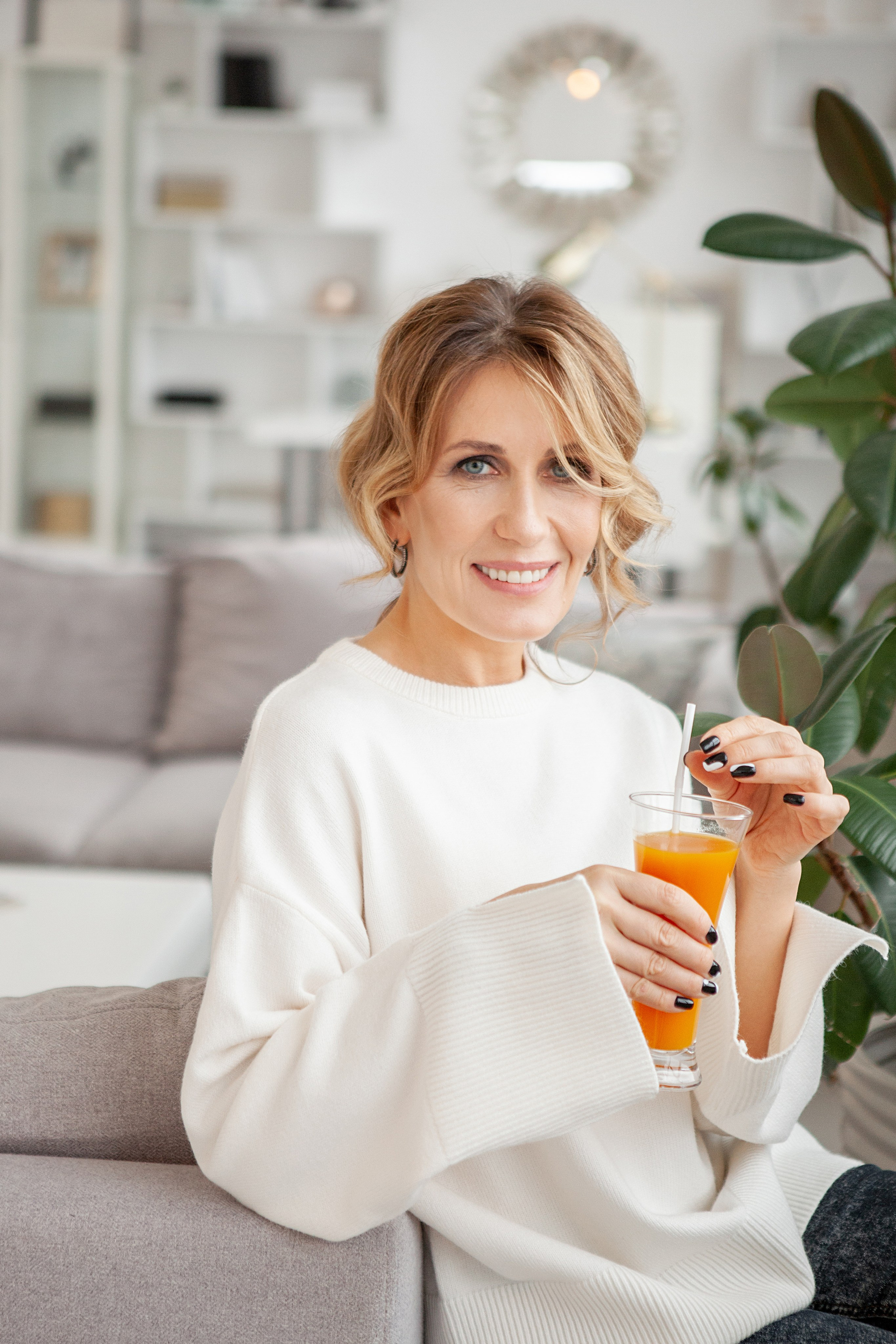 женщина в белом свитере пьет оранжевый сок и улыбается.