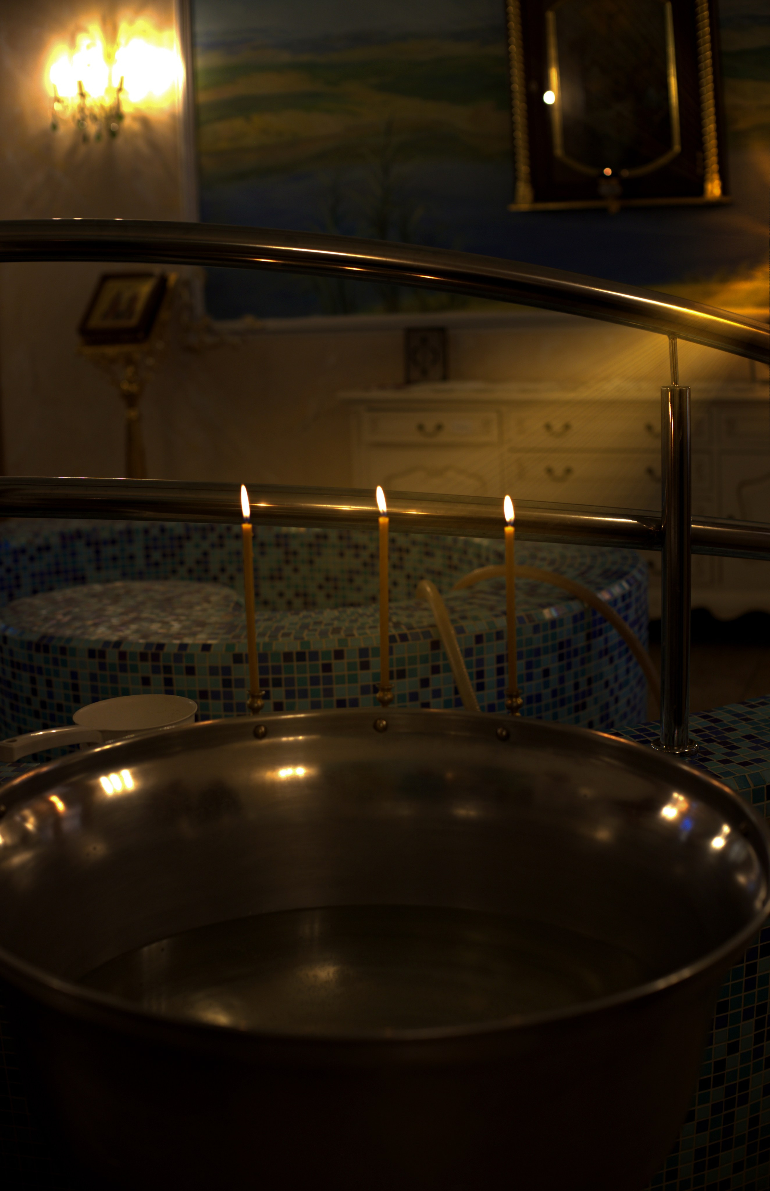 Металлическая чаша со святой водой и три горящие свечи на переднем плане, на заднем фоне мозаичная купель для крещения