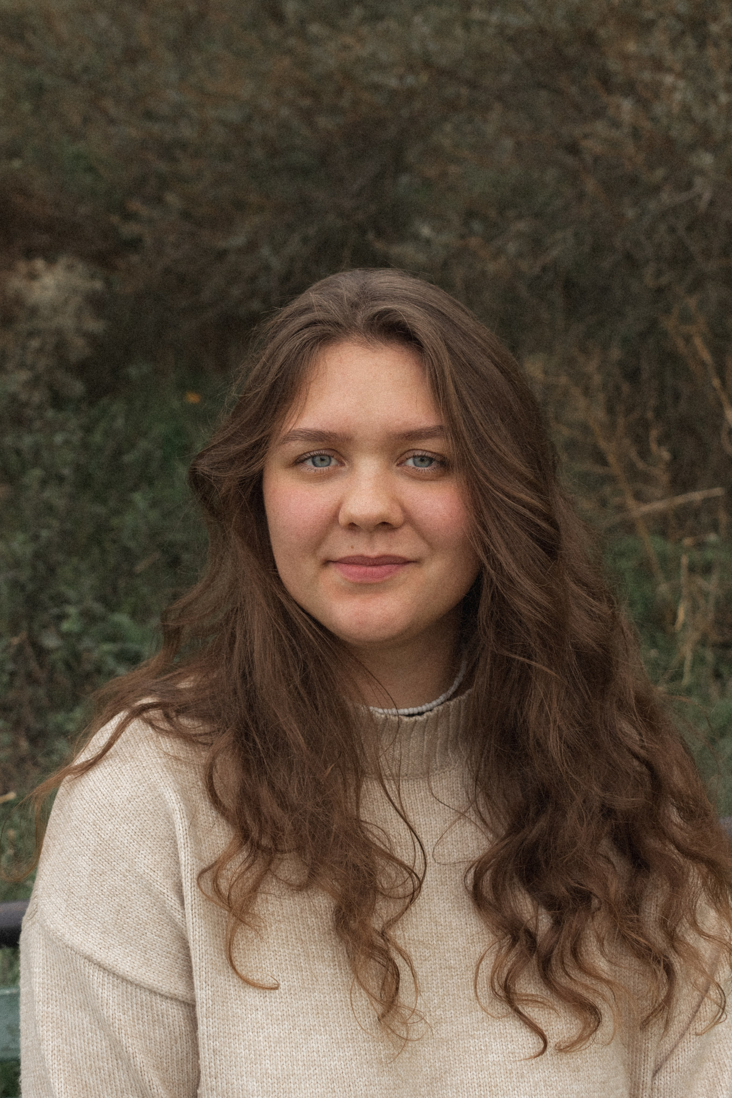 Портрет девушки в светло-бежевом свитере с каштановыми кудрявыми волосами на фоне зелени