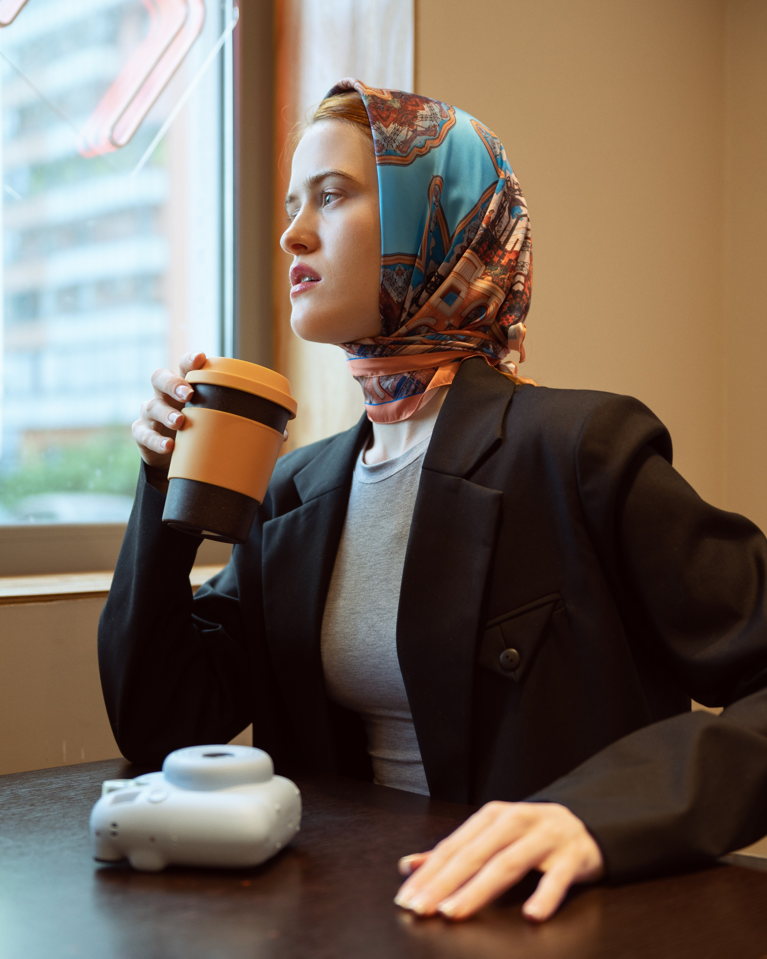 Красивая девушка в ярком платке сидит в кофейне, в руке у нее  желтый стакан для кофе, а на столе голубой Instax, 