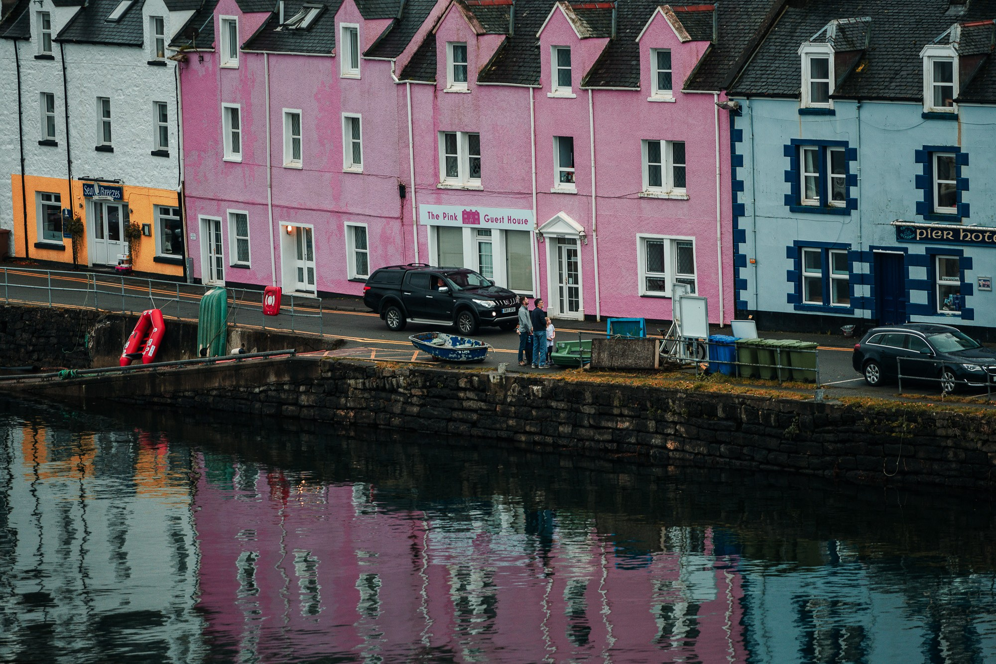 Маленькая лодка и автомобили припаркованы у причала в гавани Портри, Шотландия, с разноцветными зданиями, включая знаменитый розовый гостевой дом, отражающимися в спокойной воде.